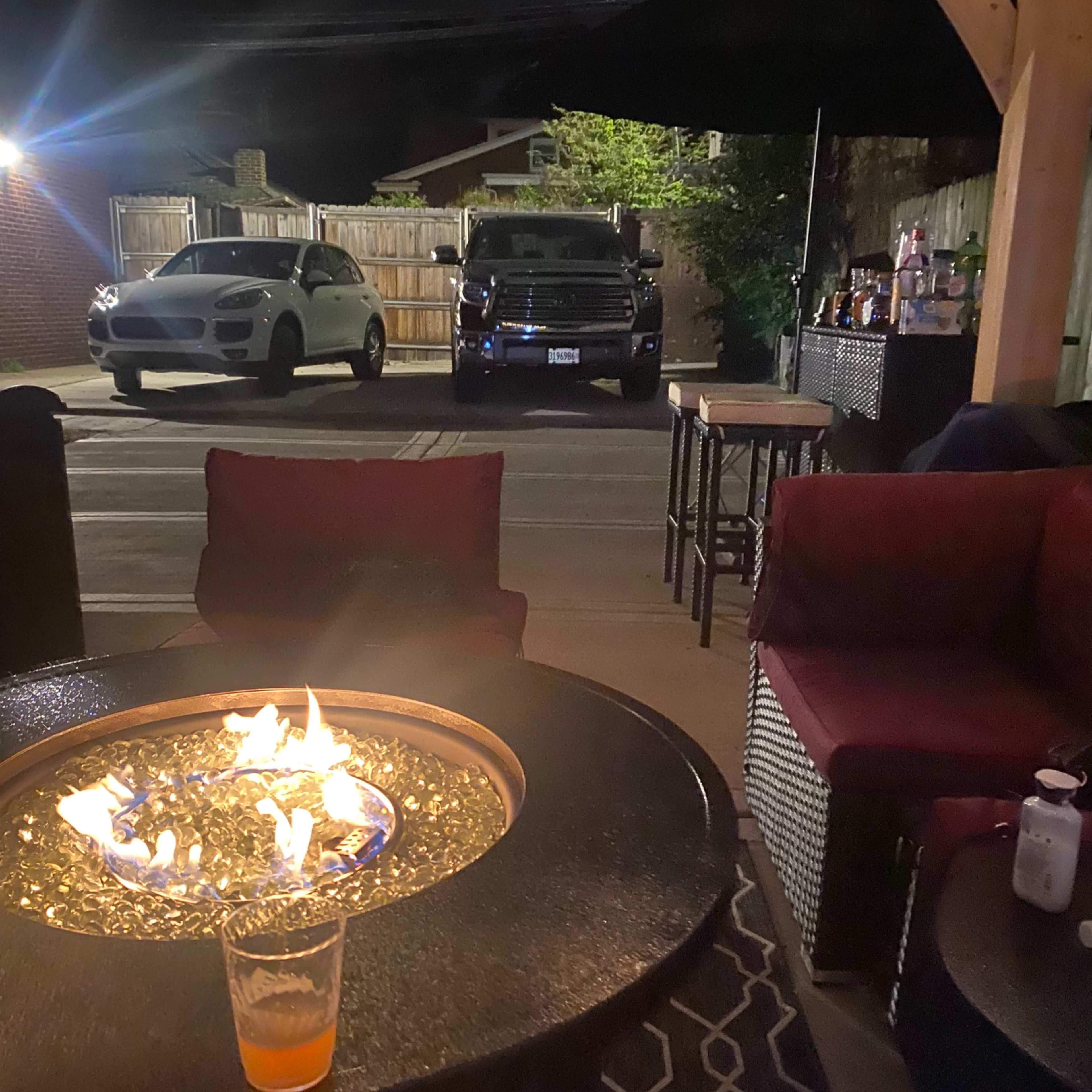 The scene shows a dimly lit outdoor area with a fire pit in the foreground, surrounded by seating, and two parked vehicles visible in the background.