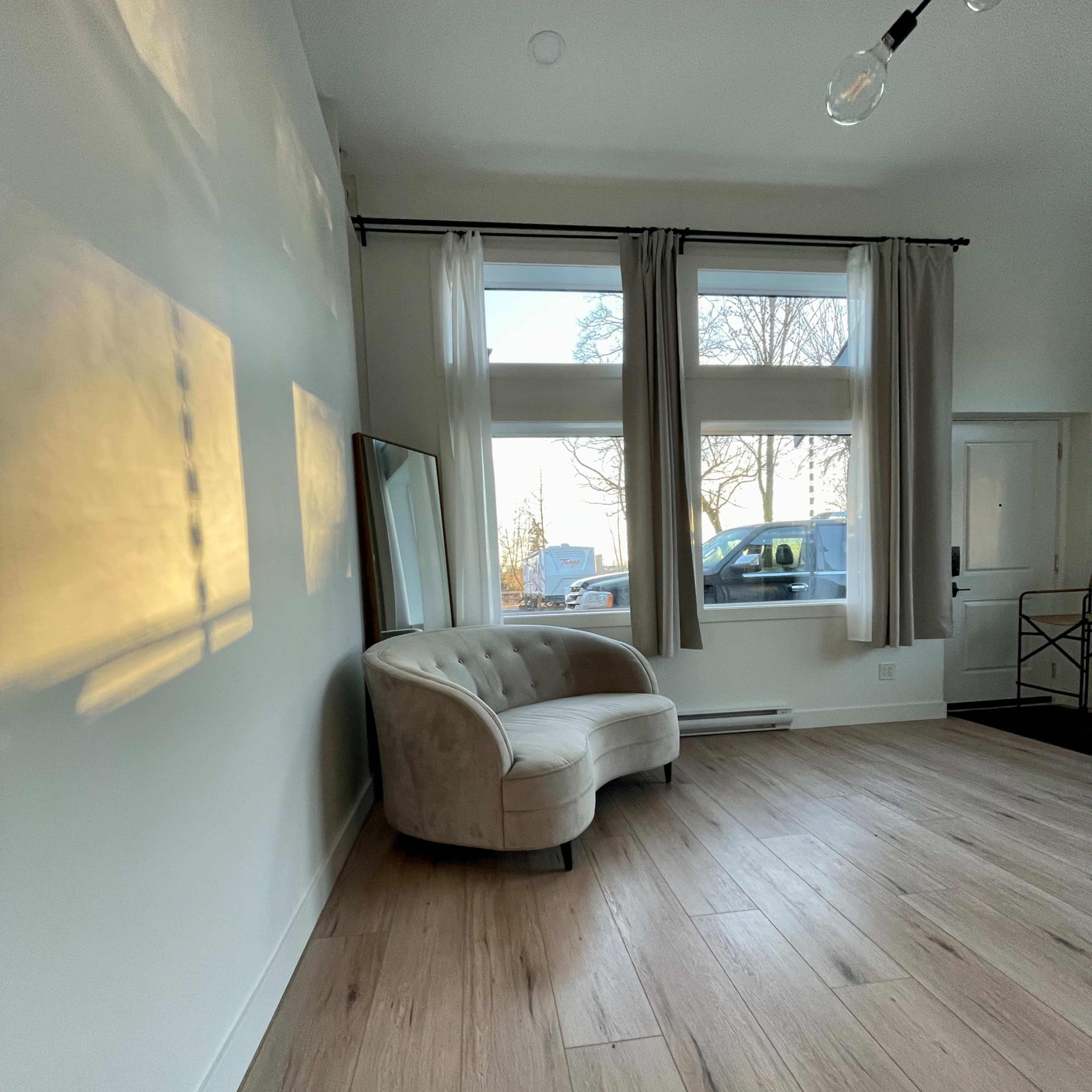 A light-colored, upholstered couch is positioned against a wall in a room with large windows and wooden flooring.