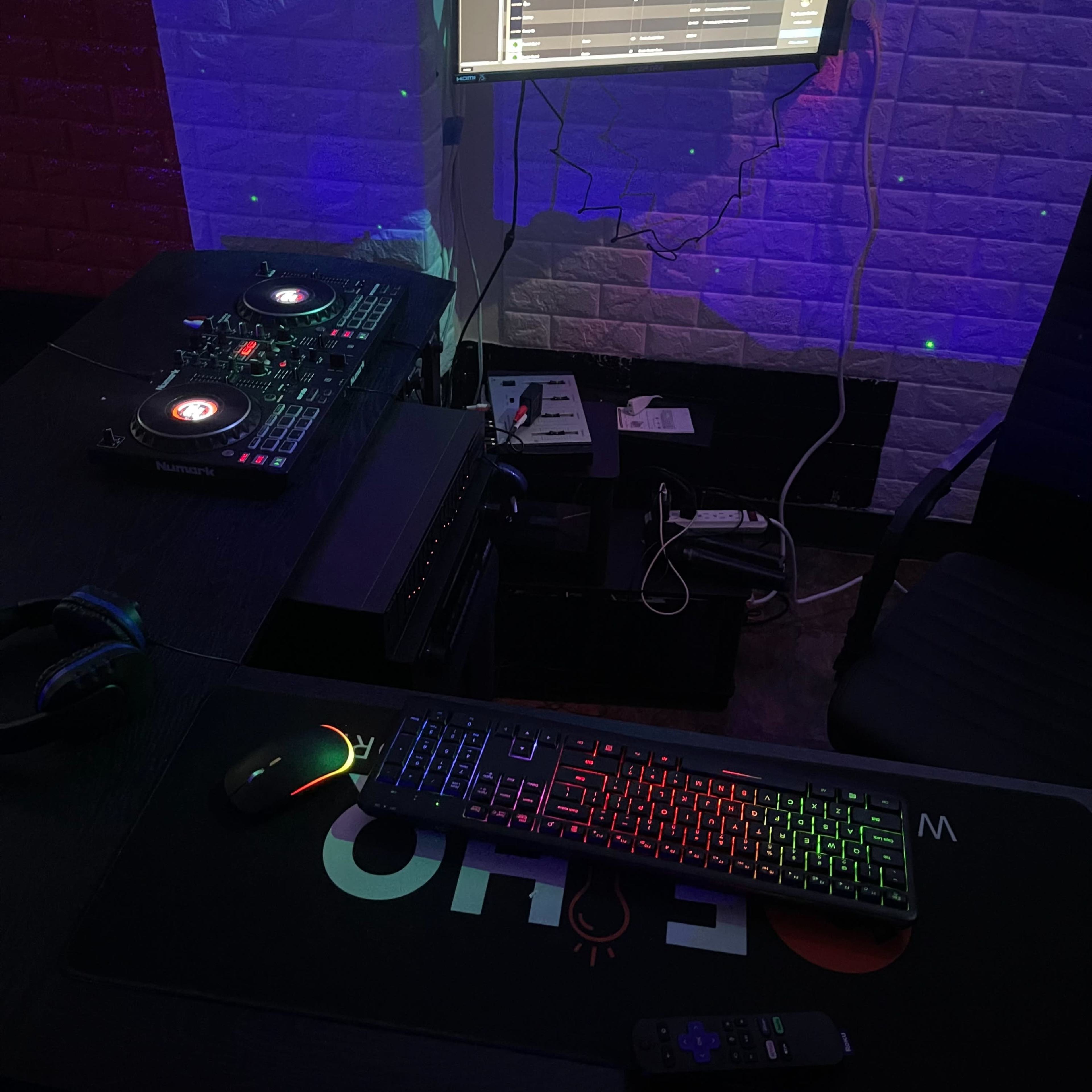 A DJ setup is displayed with turntables, a computer monitor showing music software, and a keyboard on a dark table beneath colorful lighting.