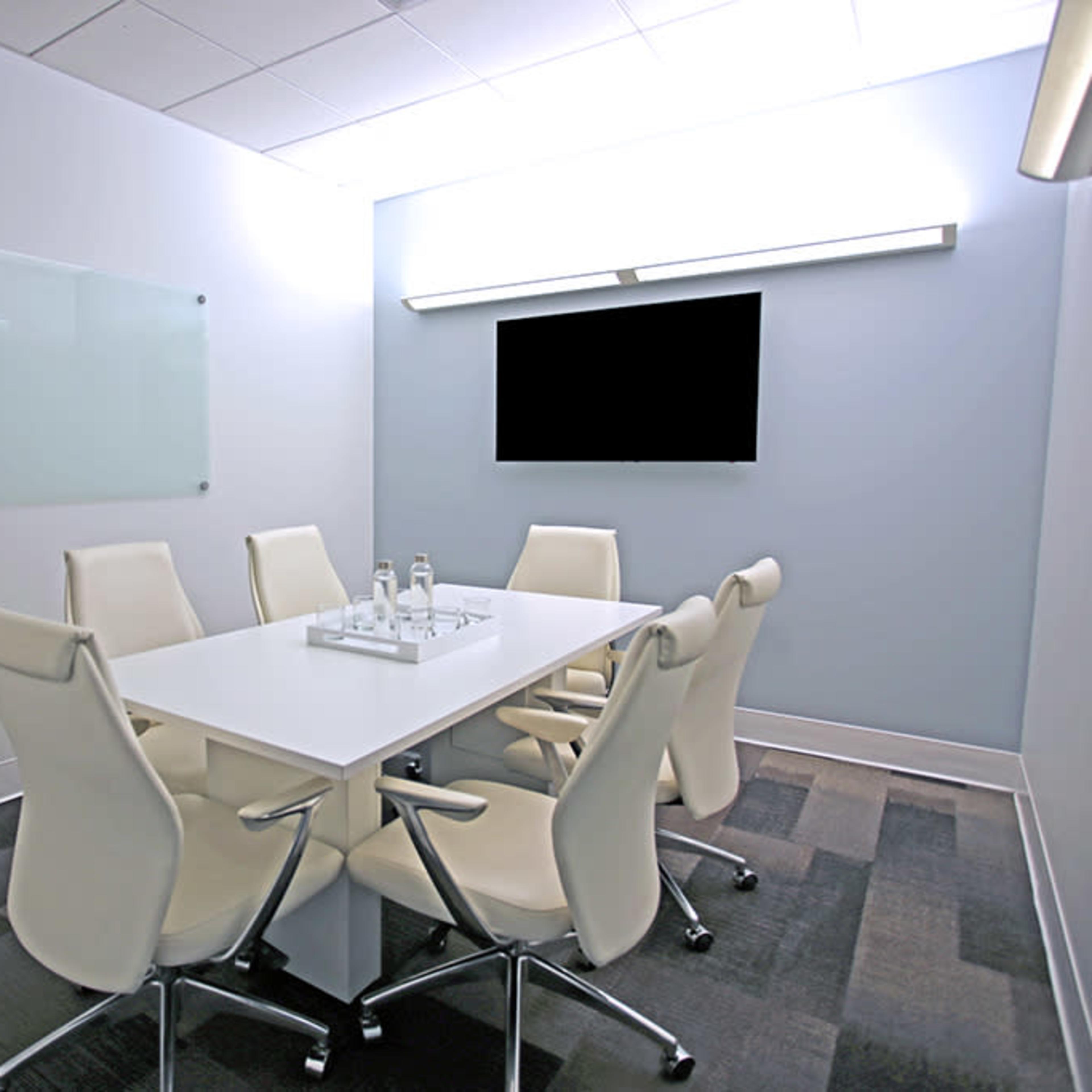 A modern conference room features a white table surrounded by six white chairs, with a glass whiteboard and a wall-mounted television.