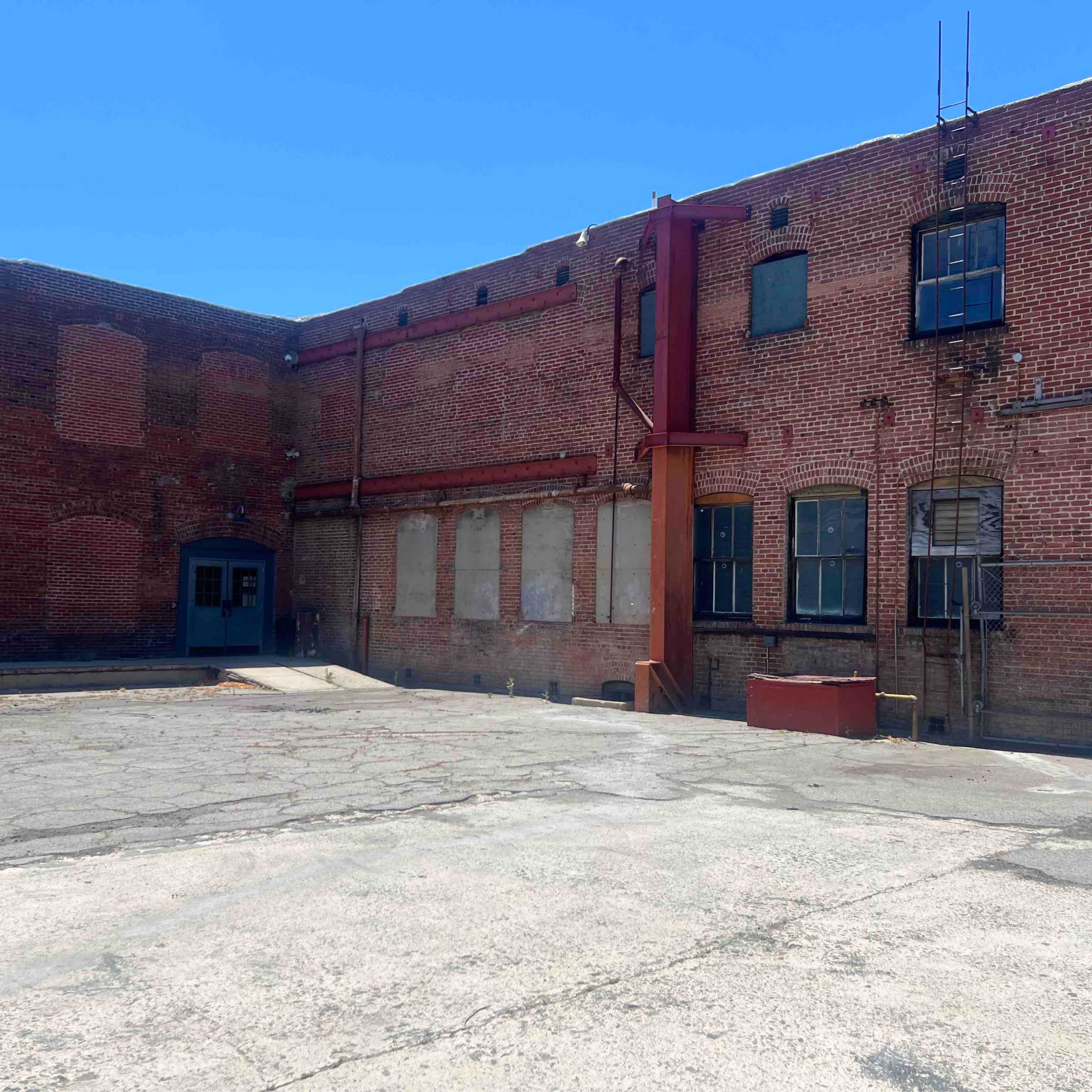 A weathered brick building with multiple windows and a large, empty concrete area in front.