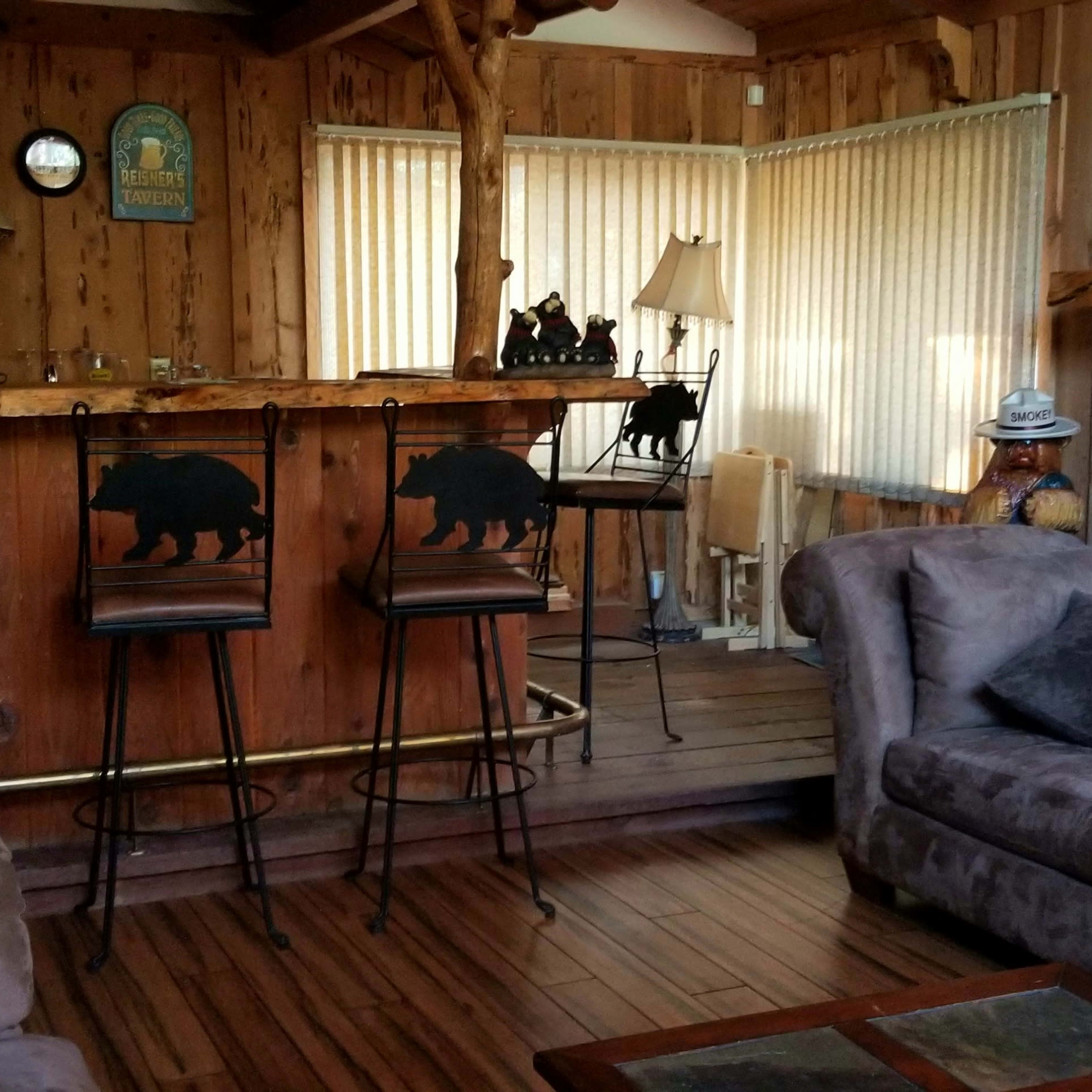 A rustic wooden interior featuring a bar with bear-themed stools, a living area with a sectional couch, and large windows covered with beige curtains.
