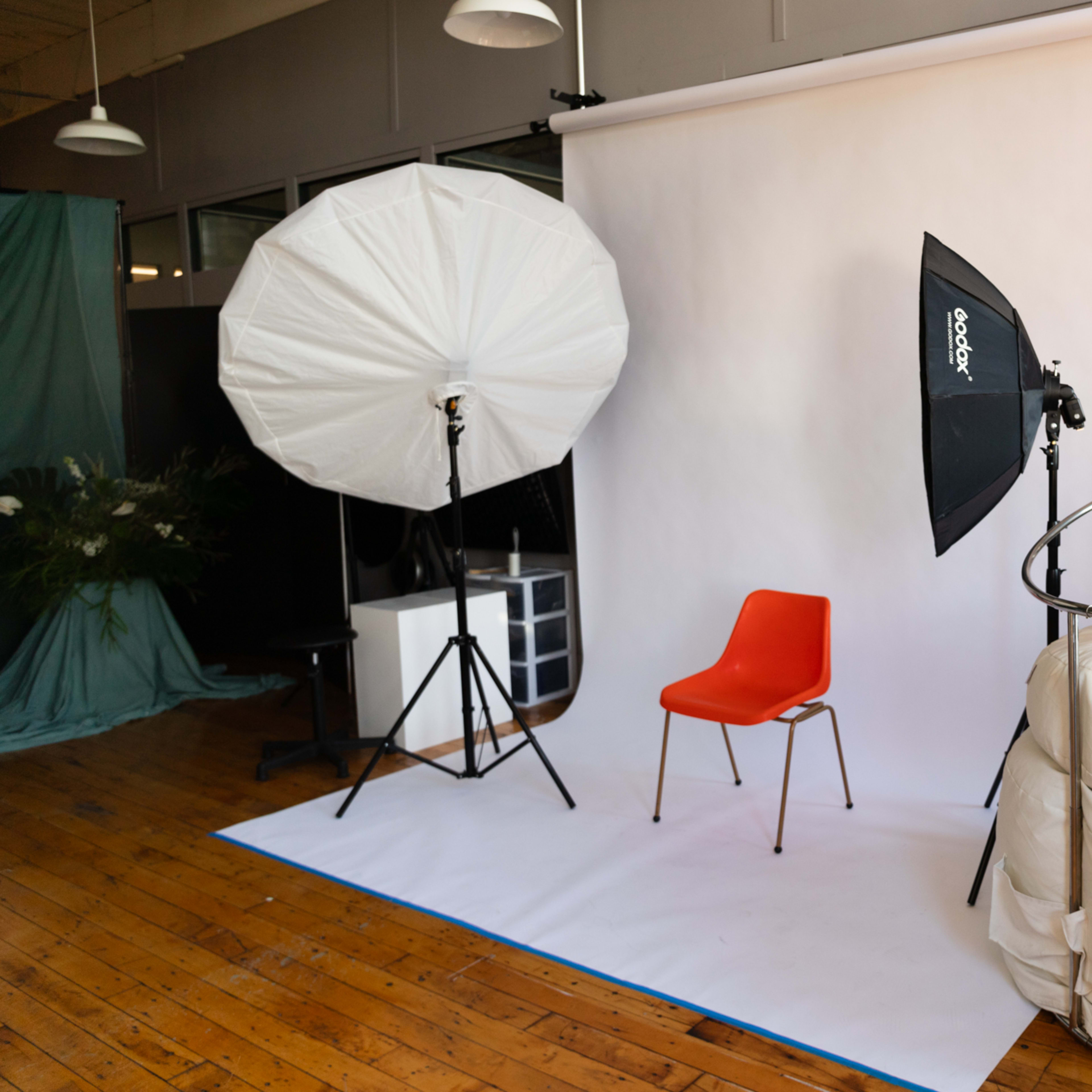 A photo studio setup features a single orange chair against a white backdrop, flanked by large softboxes and a light umbrella.