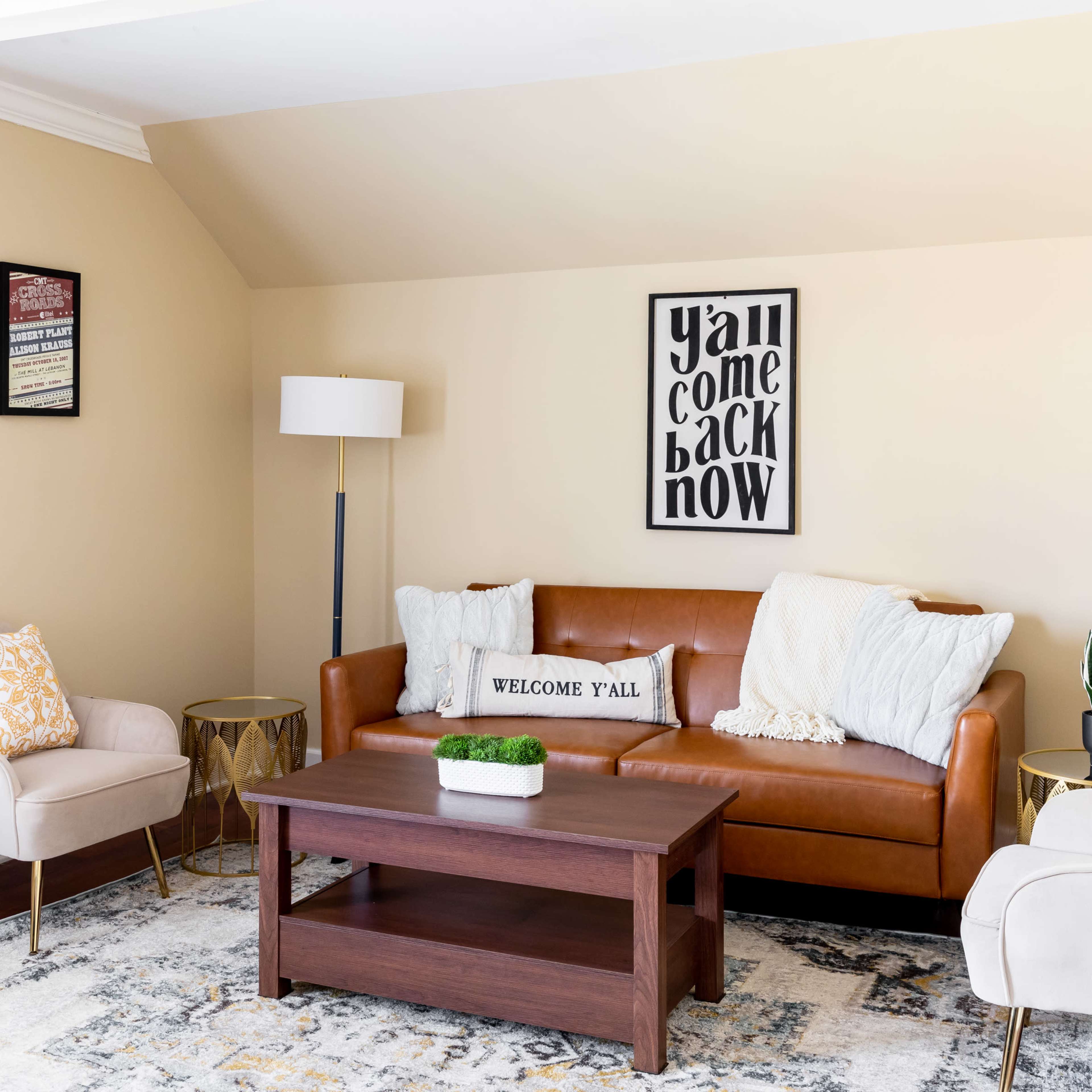A cozy living room features a brown leather sofa, two light-colored armchairs, a wooden coffee table, and a decorative wall sign.
