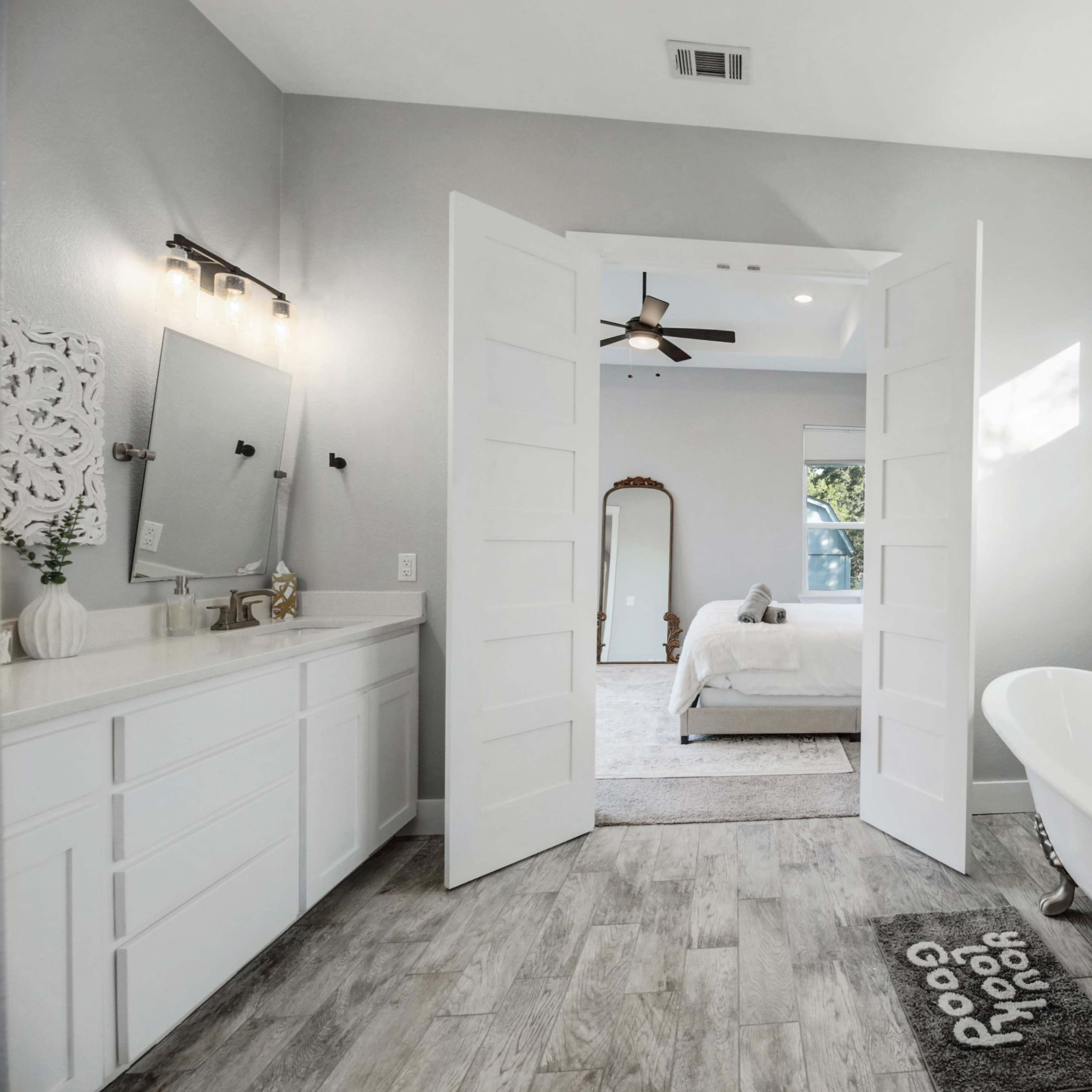 A spacious bathroom features a freestanding bathtub, double vanity, and an open doorway leading to a well-lit bedroom.