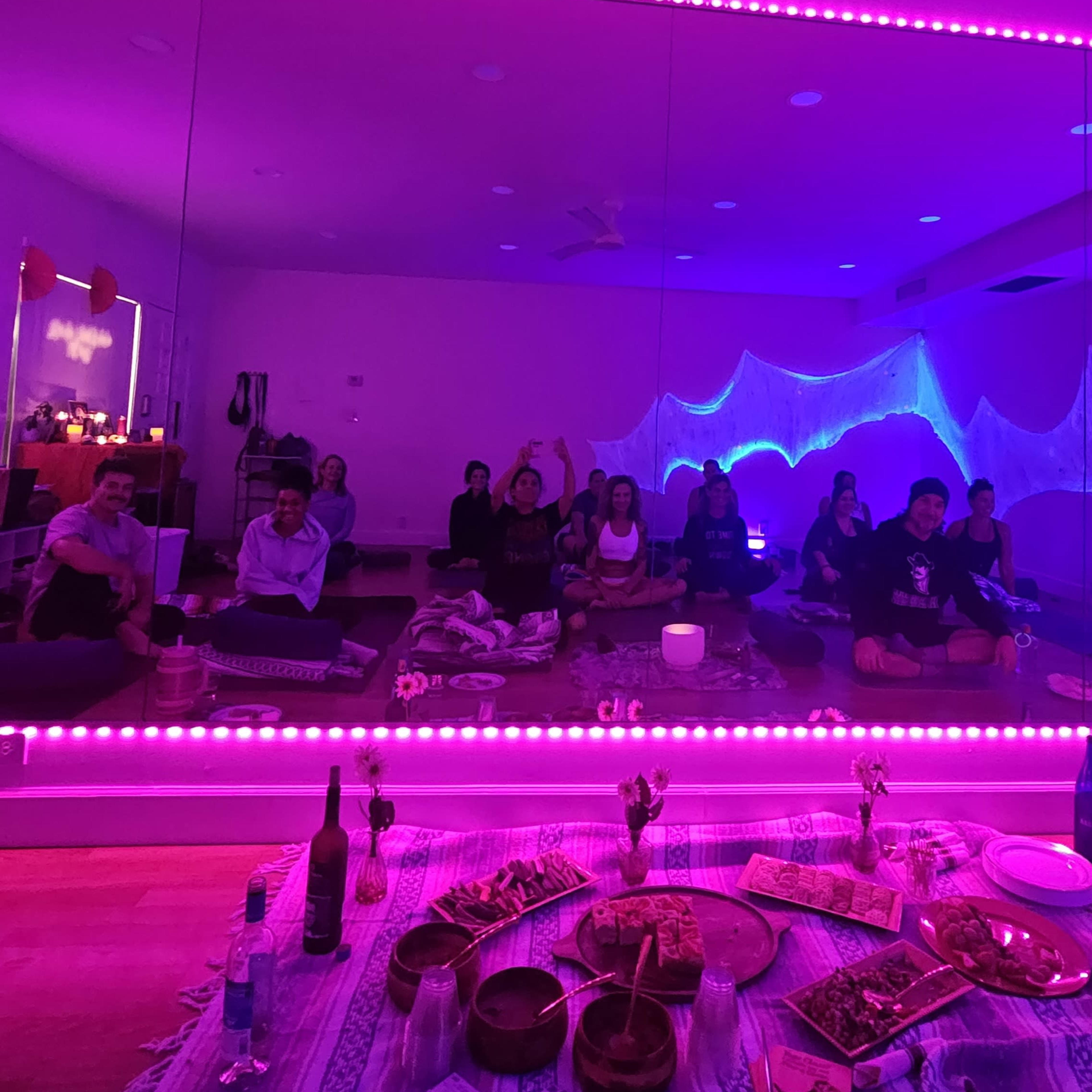A group of people sits on the floor in a brightly lit room with purple ambient lighting, reflected in a large mirror, while food is displayed on a central table.