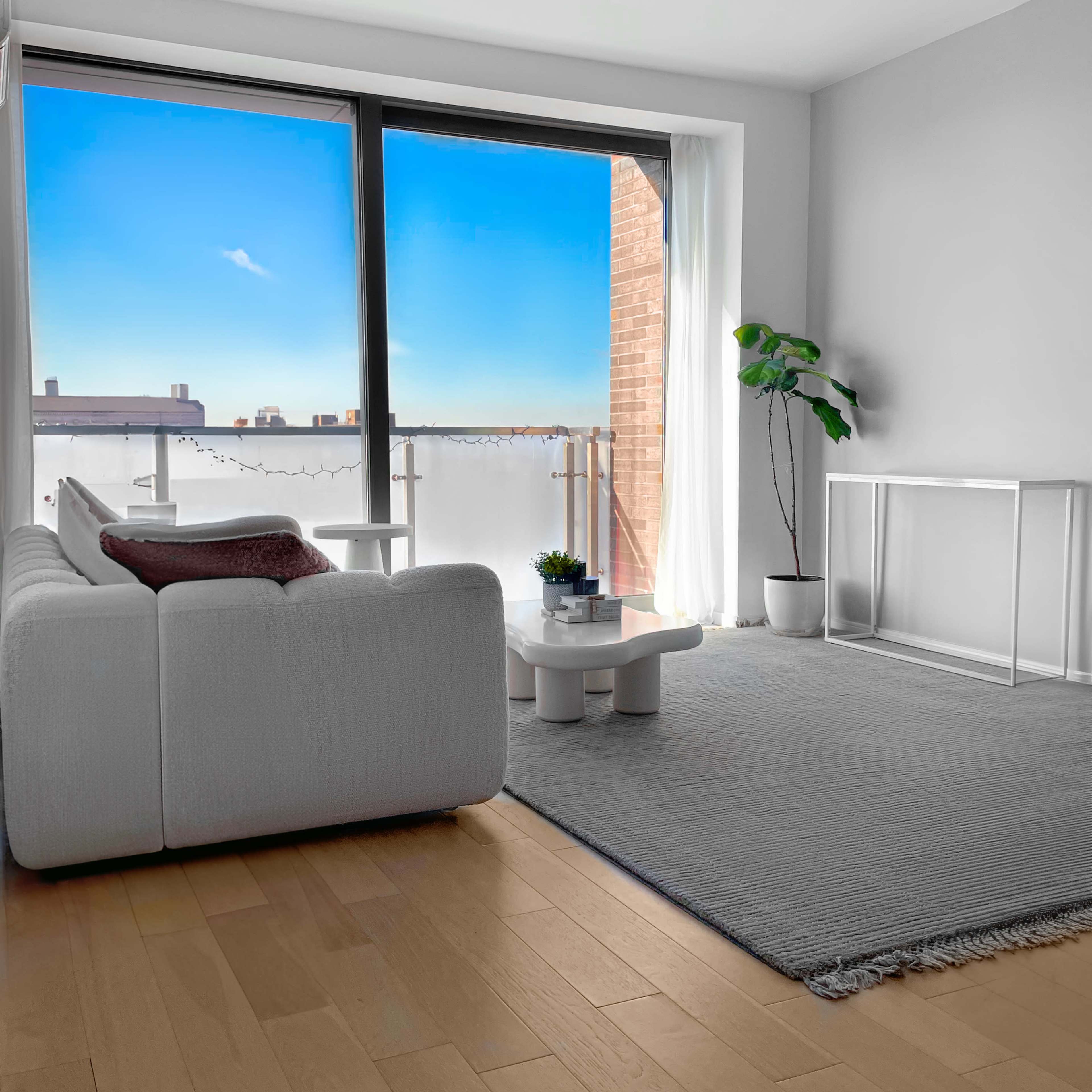 A modern living room with a white sofa, a coffee table, a rug, and large windows illuminating the space with a view of a blue sky.
