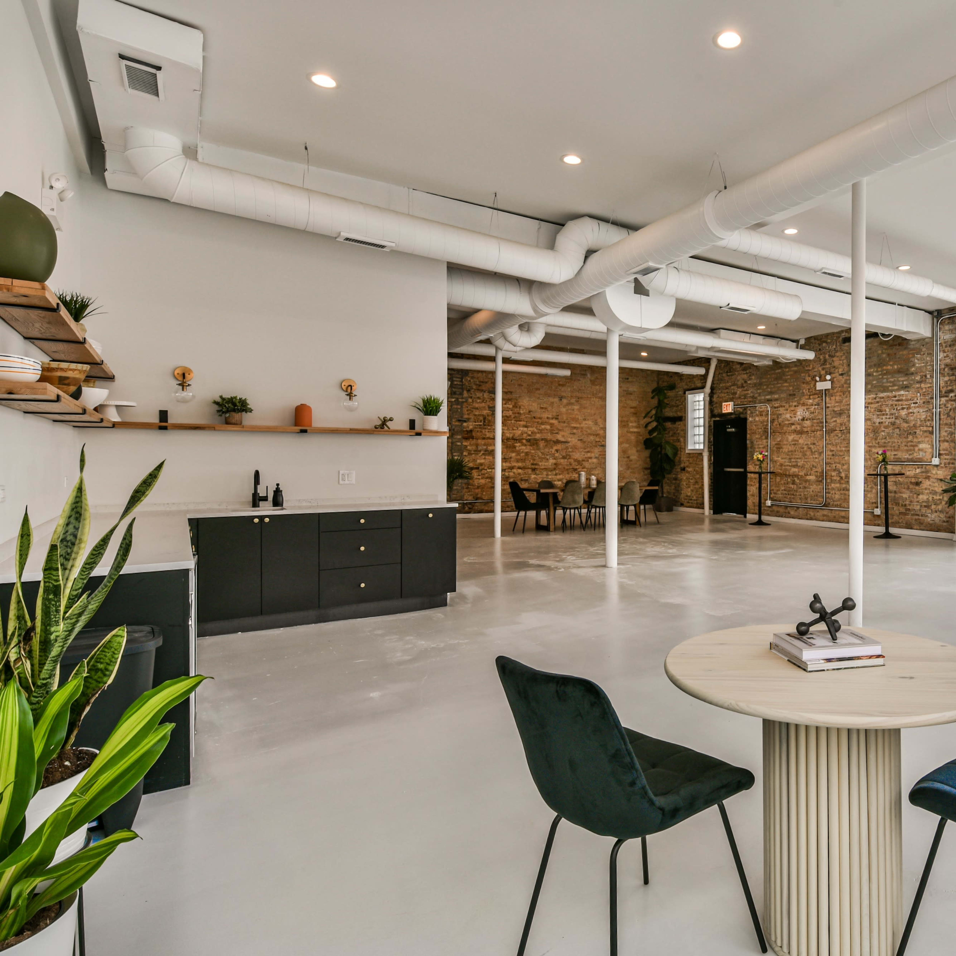 A spacious, modern interior featuring a round table with two chairs, exposed brick walls, and large windows allowing natural light.