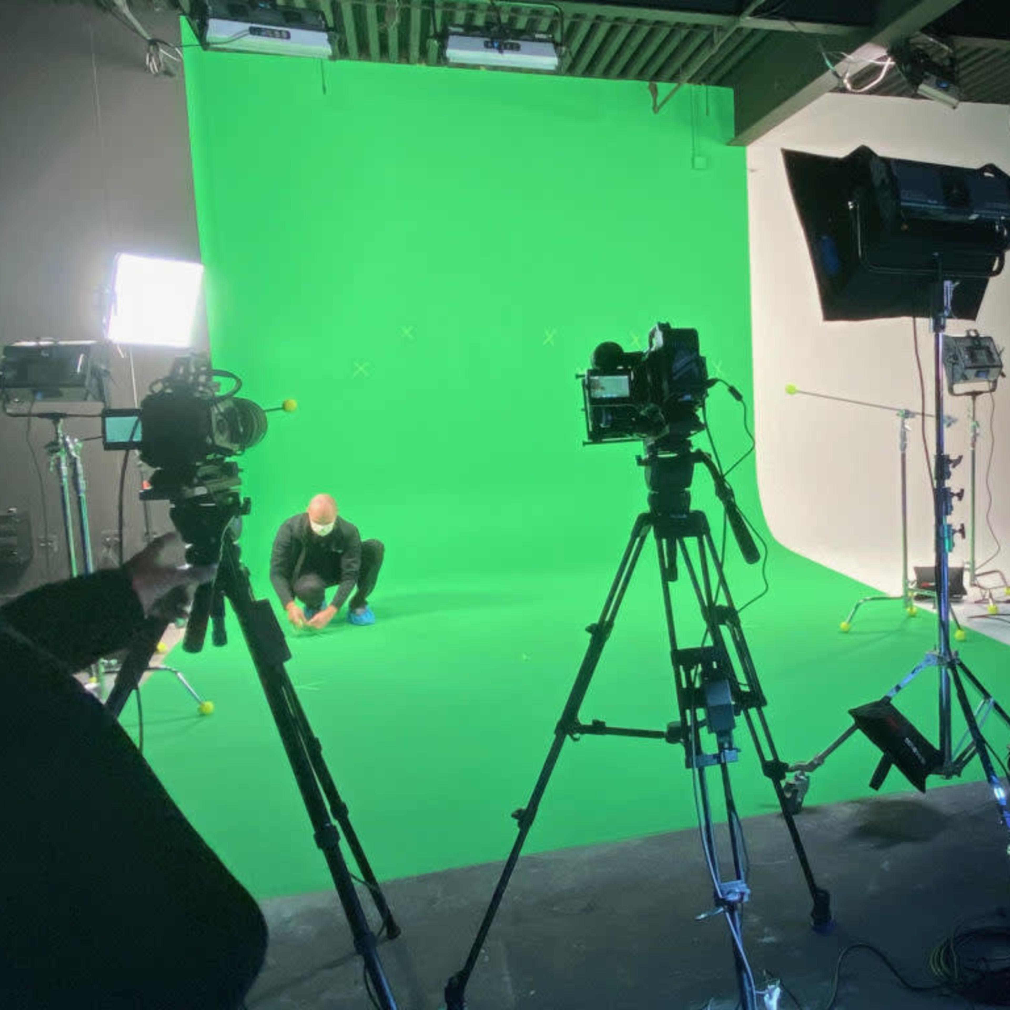 Two individuals are setting up equipment in a studio with a green screen backdrop, surrounded by cameras and lighting.