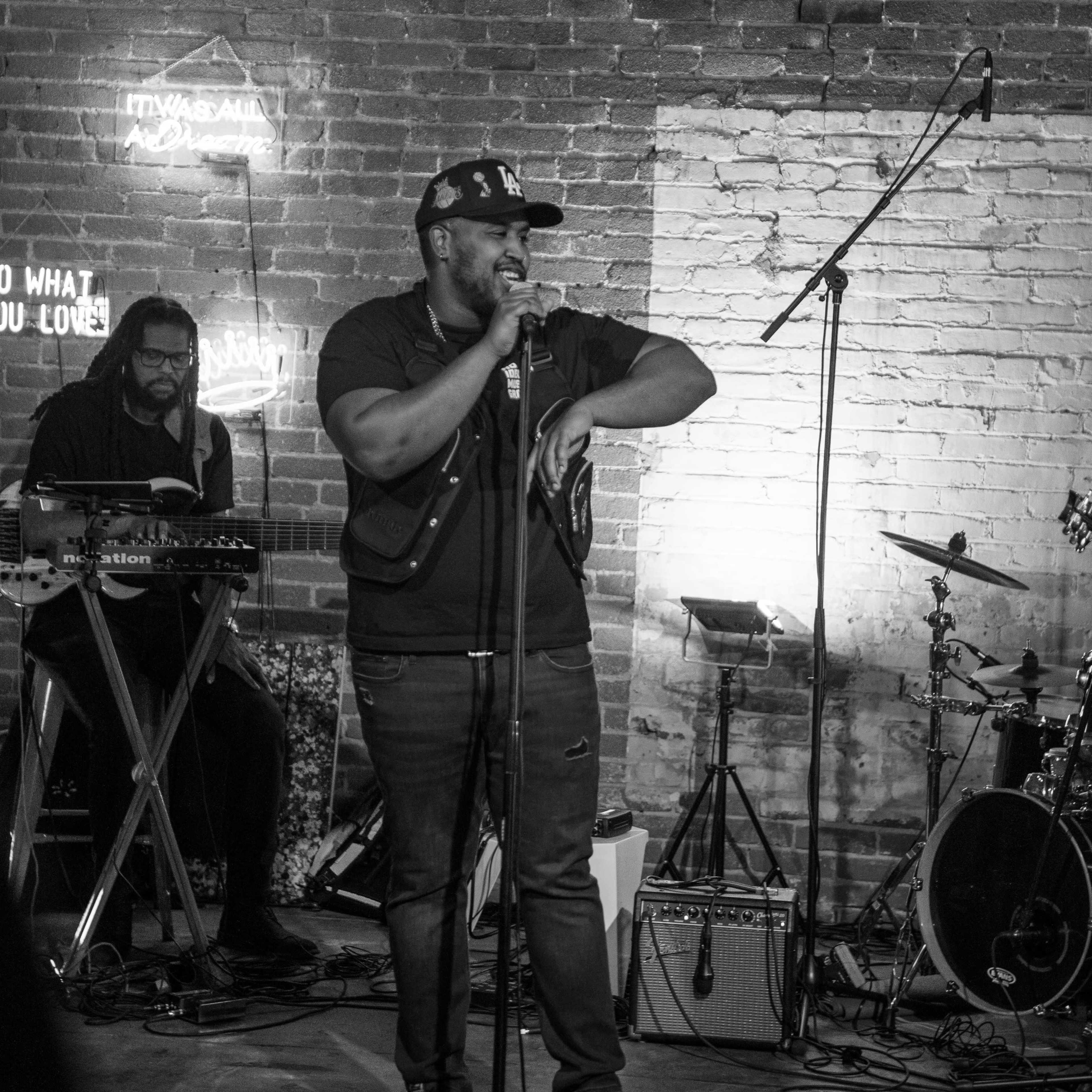 A man stands on stage singing into a microphone while another musician plays a keyboard in the background.