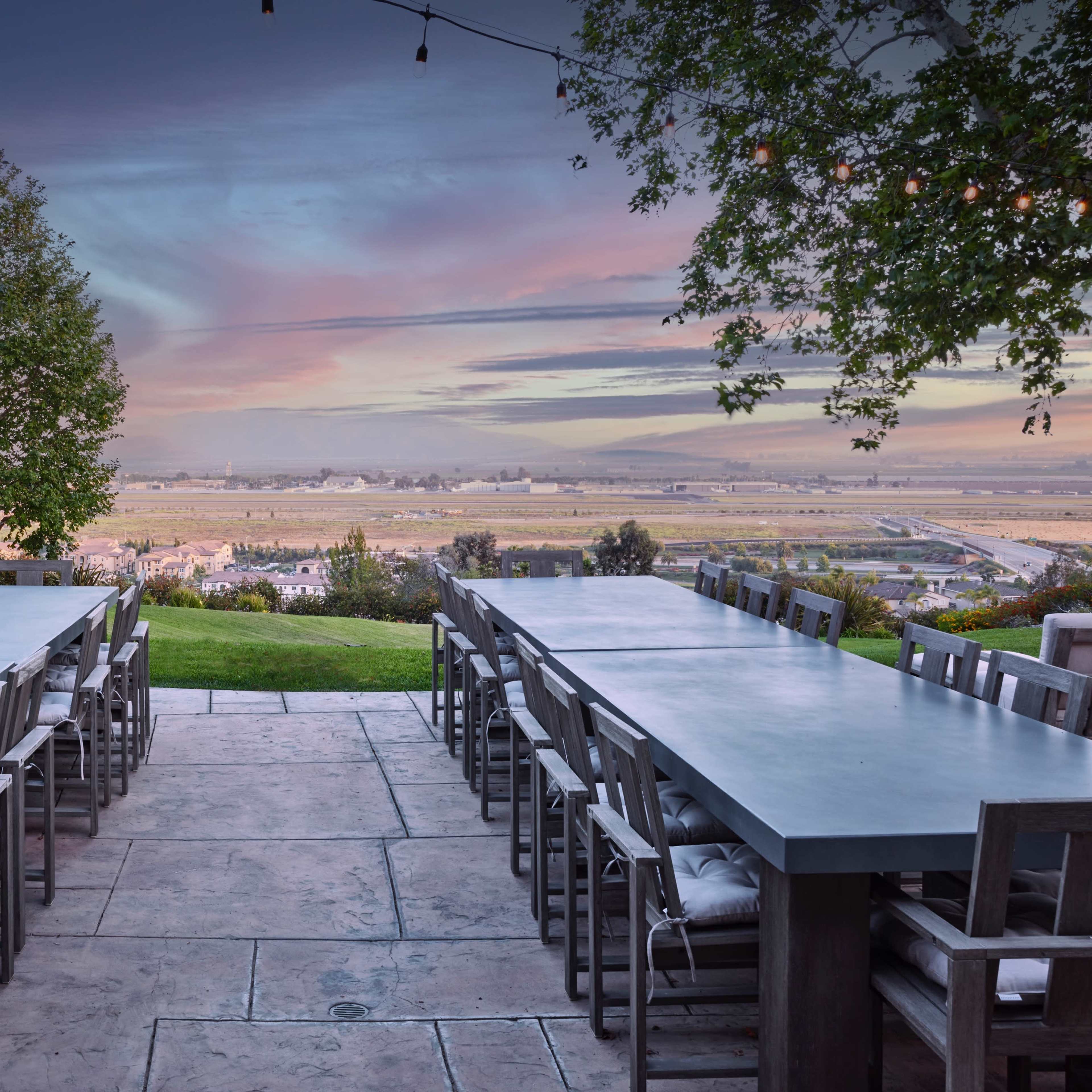The scene features an outdoor dining area with long tables and chairs overlooking a valley at sunset.
