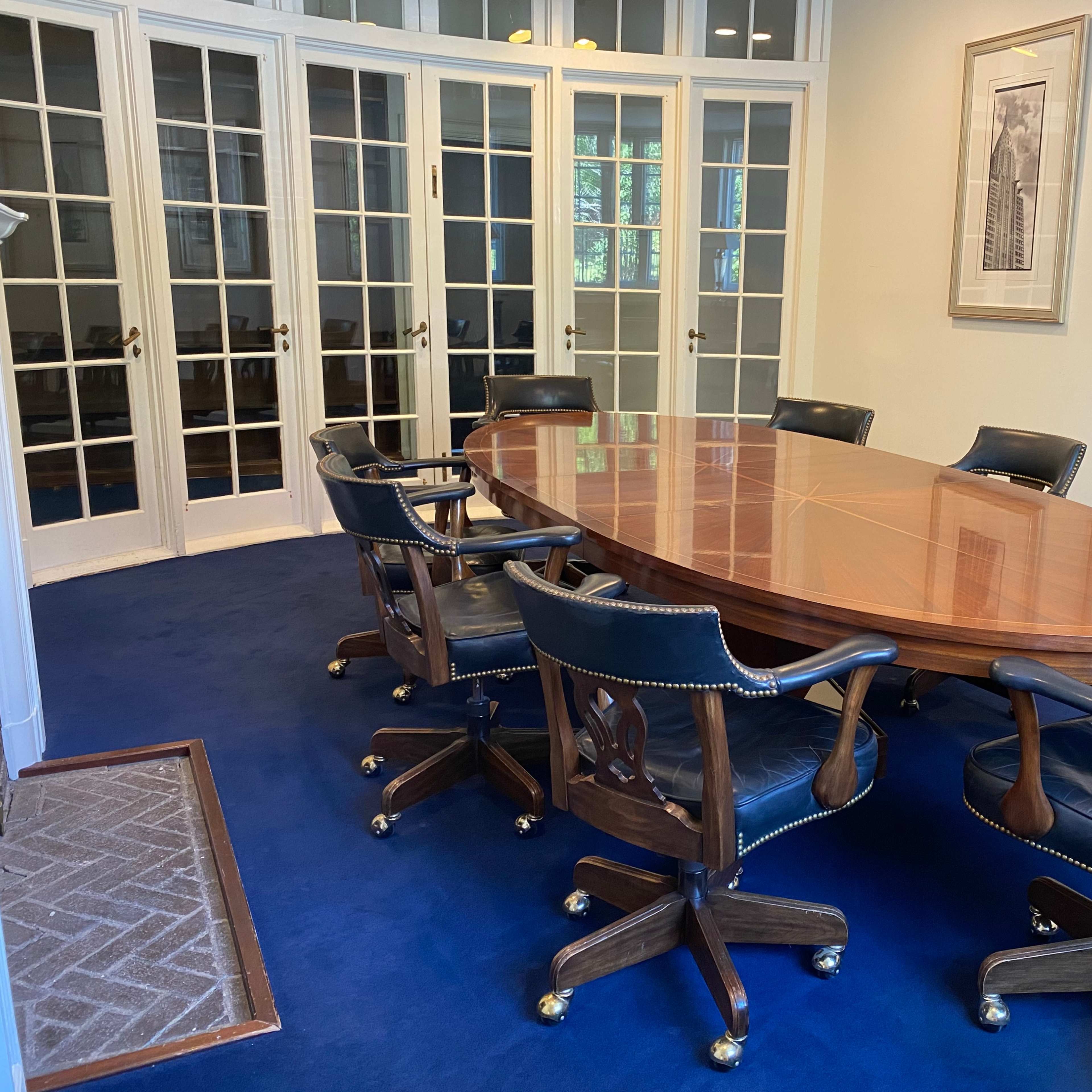 A polished wooden conference table surrounded by upholstered swivel chairs is set in a well-lit meeting room with glass-paneled doors and framed art on the walls.