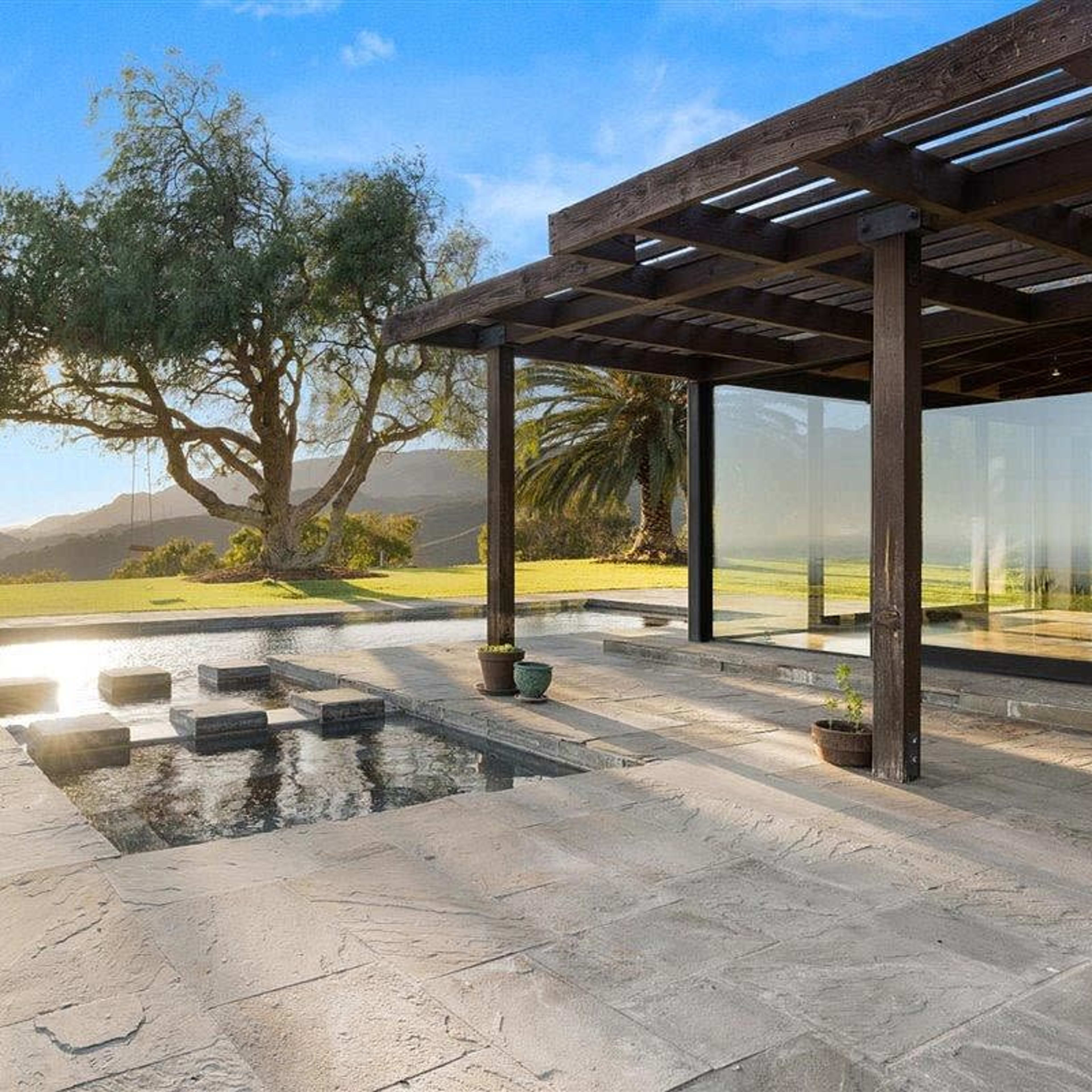 A modern home exterior with a patio, stepping stones leading to a small pool, and a large tree in the background.