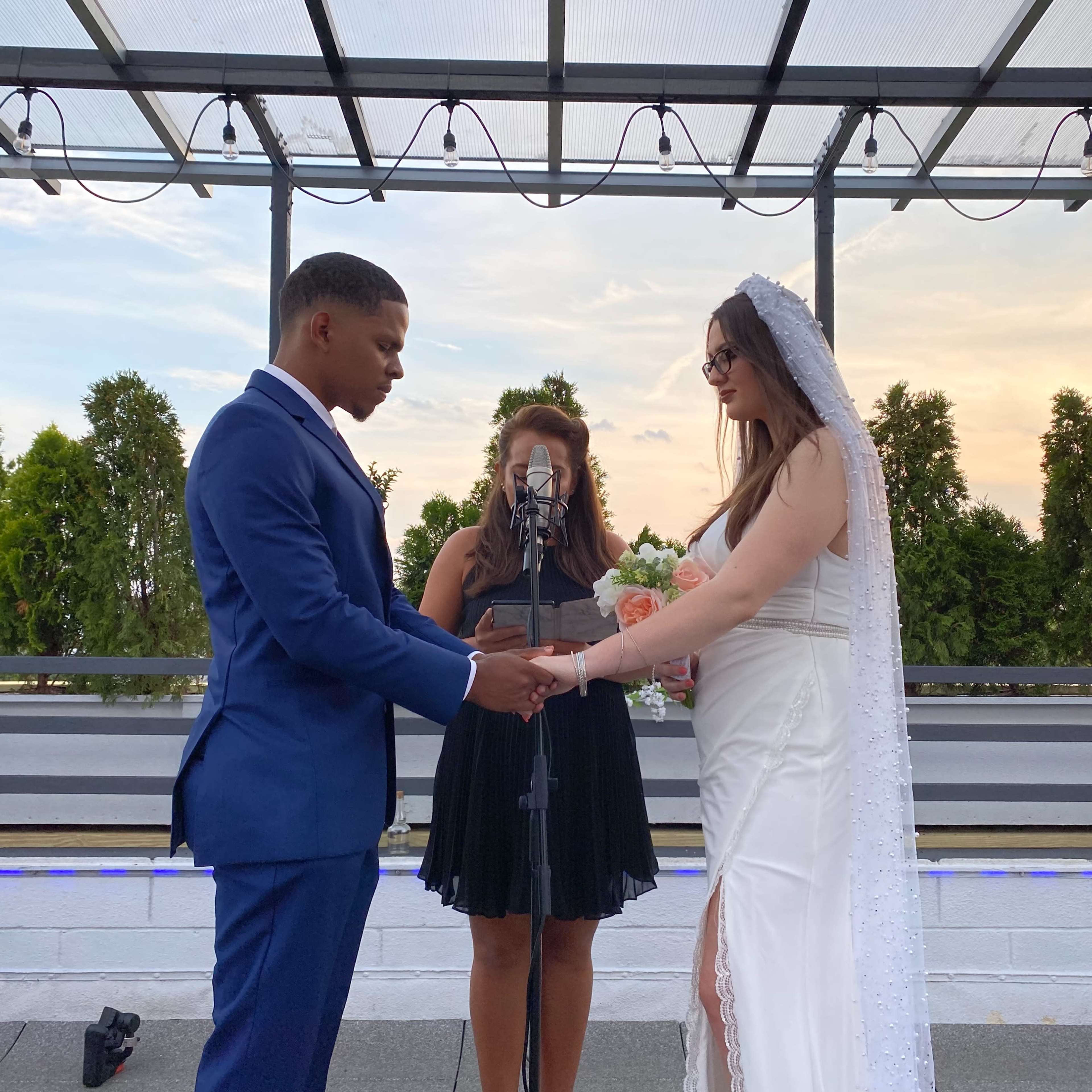 A couple stands holding hands during a wedding ceremony on a covered outdoor stage.