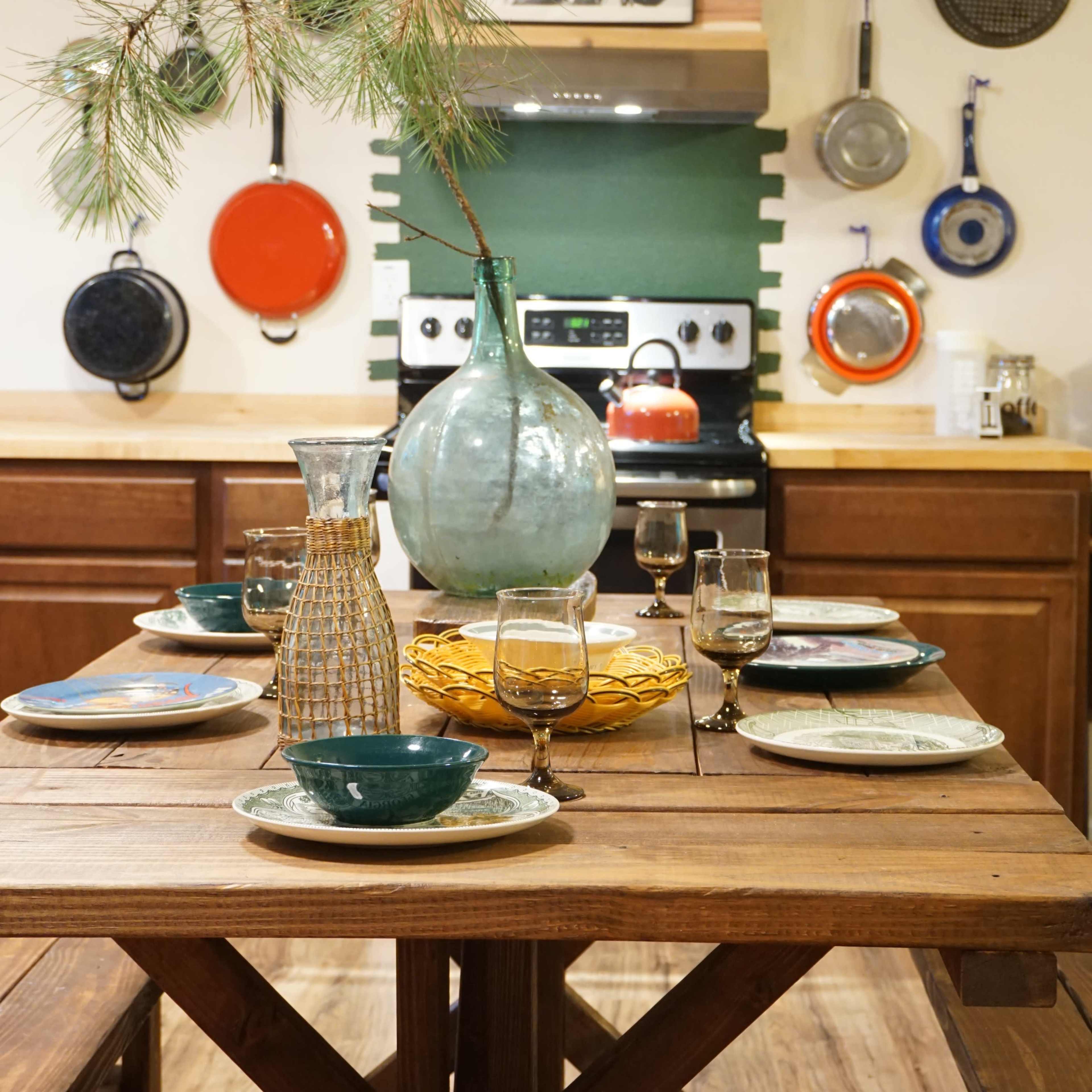 A wooden dining table is set with plates and glasses, surrounded by a kitchen featuring colorful pots and an image of a soldier on the wall.