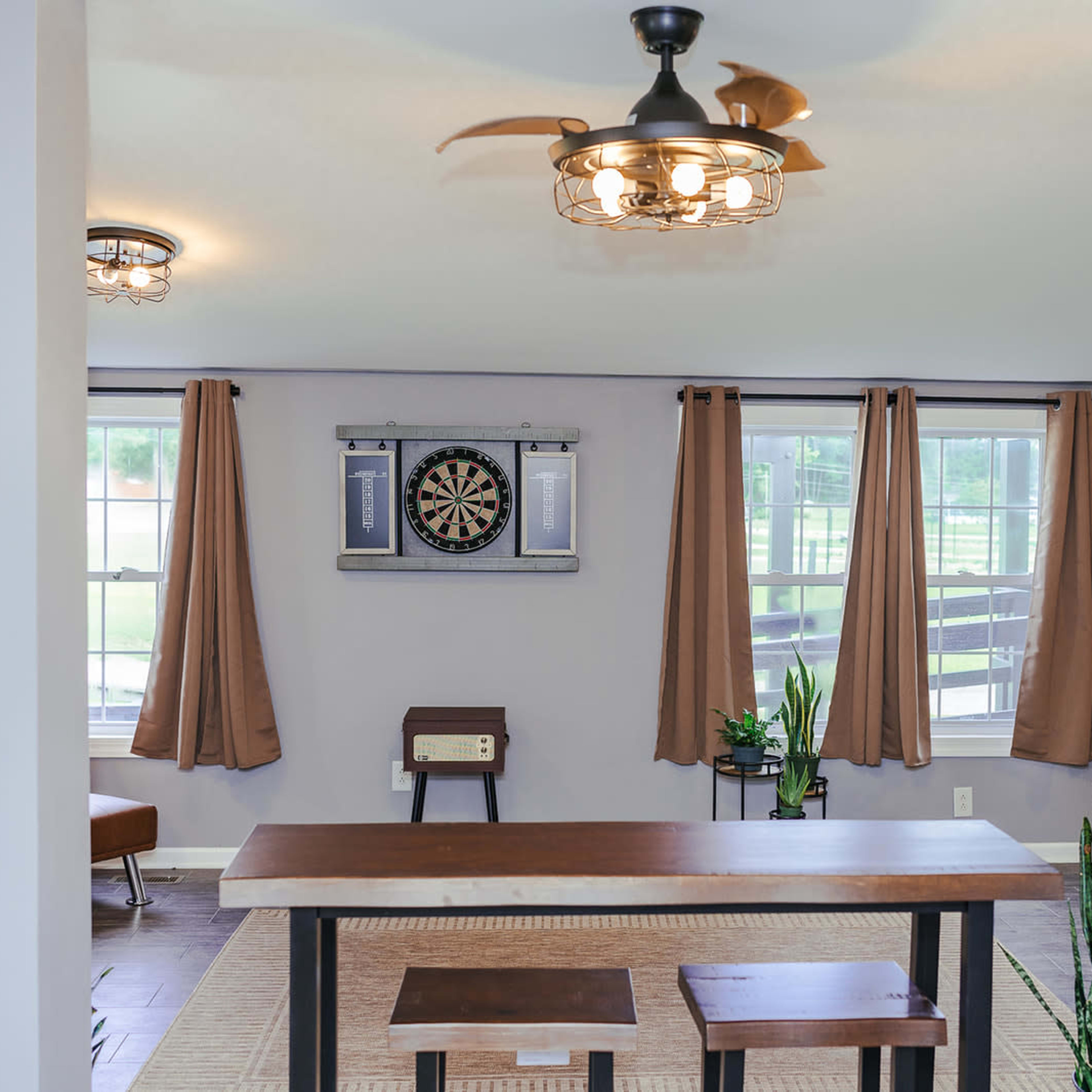 A spacious room with a wooden table and two stools in the center, a dartboard mounted on the wall, and large windows with curtain drapes.