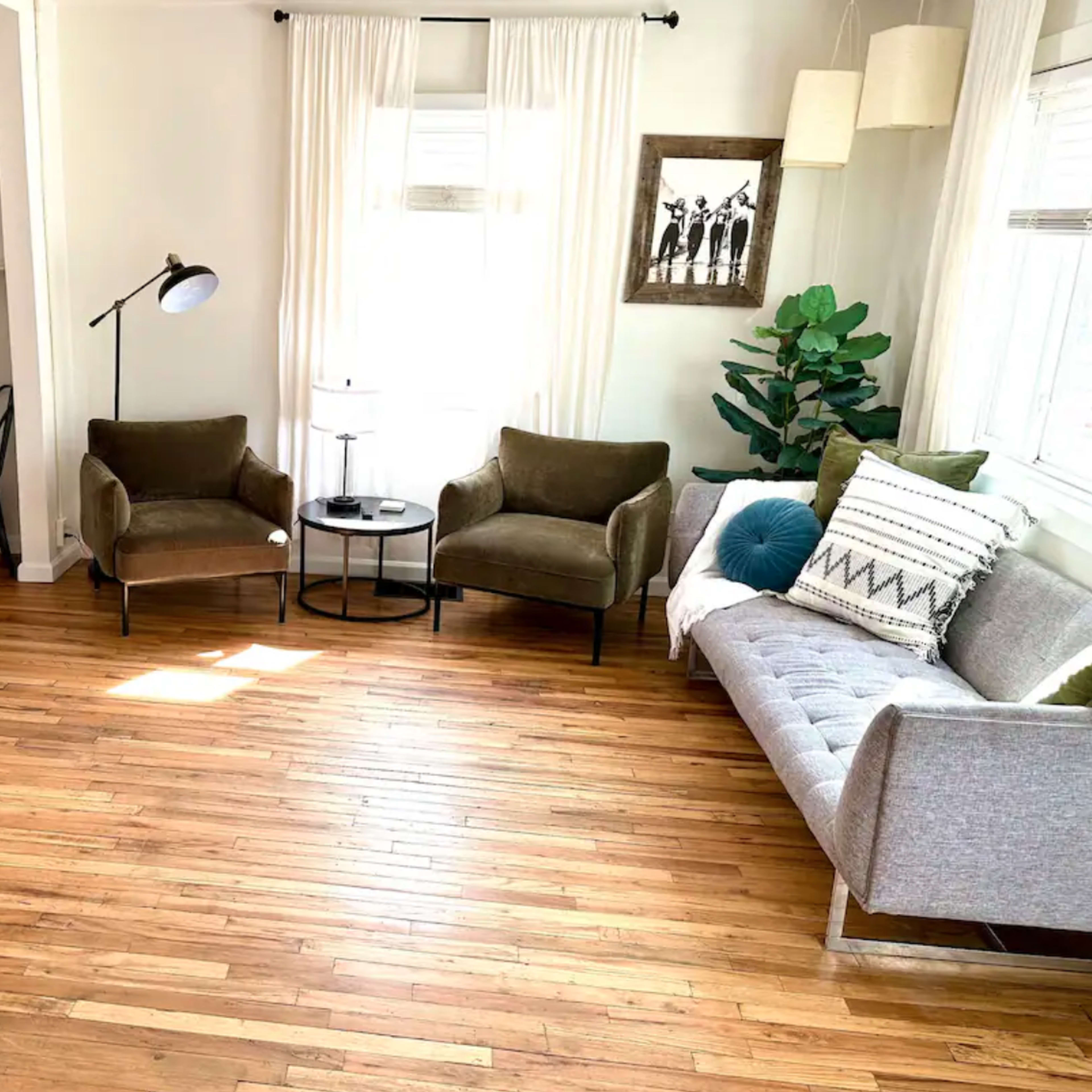 A modern living room features a gray sofa, two green armchairs, a round coffee table, a flat-screen TV on the wall, and large windows with white curtains.