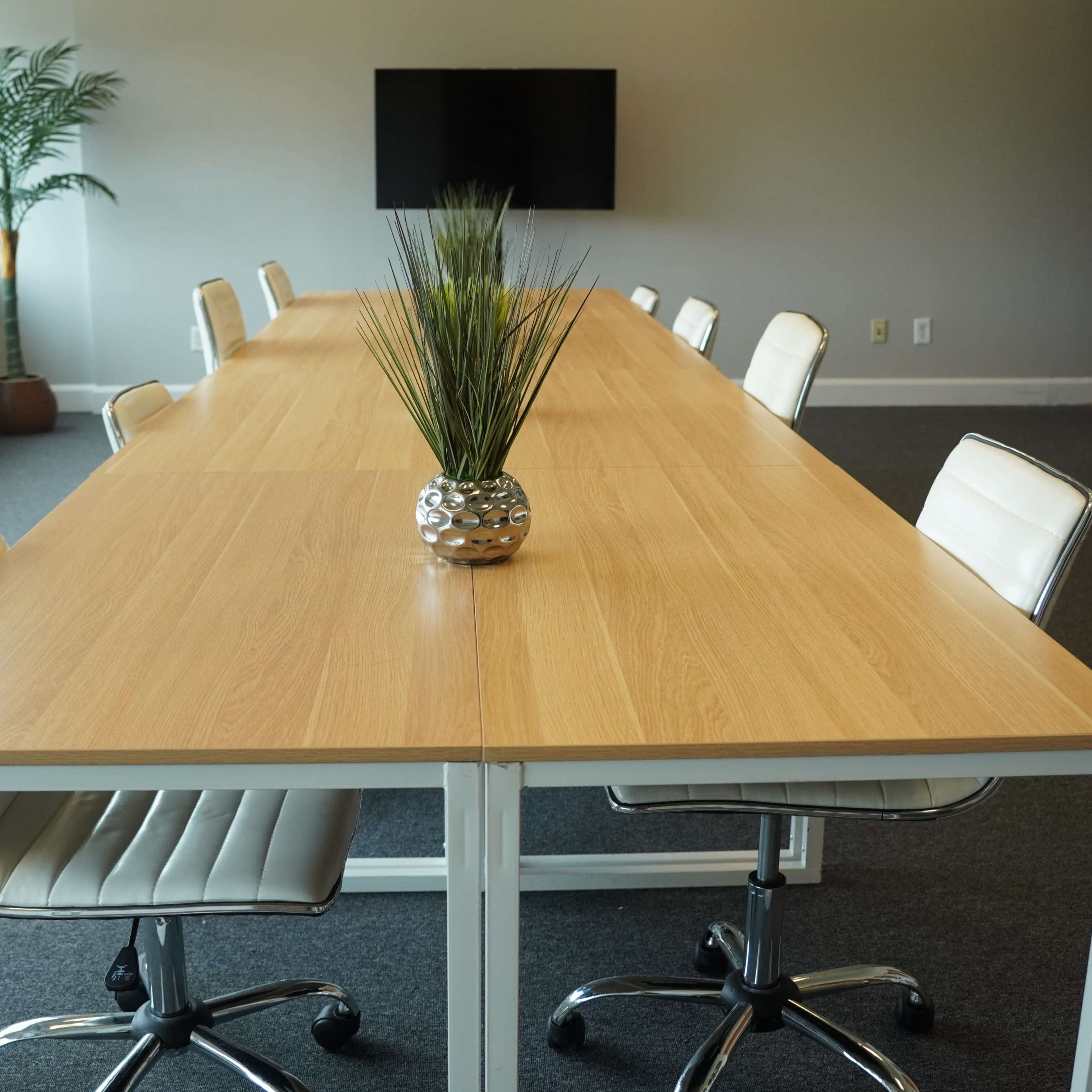 A long wooden conference table with white chairs is set in a well-lit meeting room featuring a large window and a television screen on the wall.