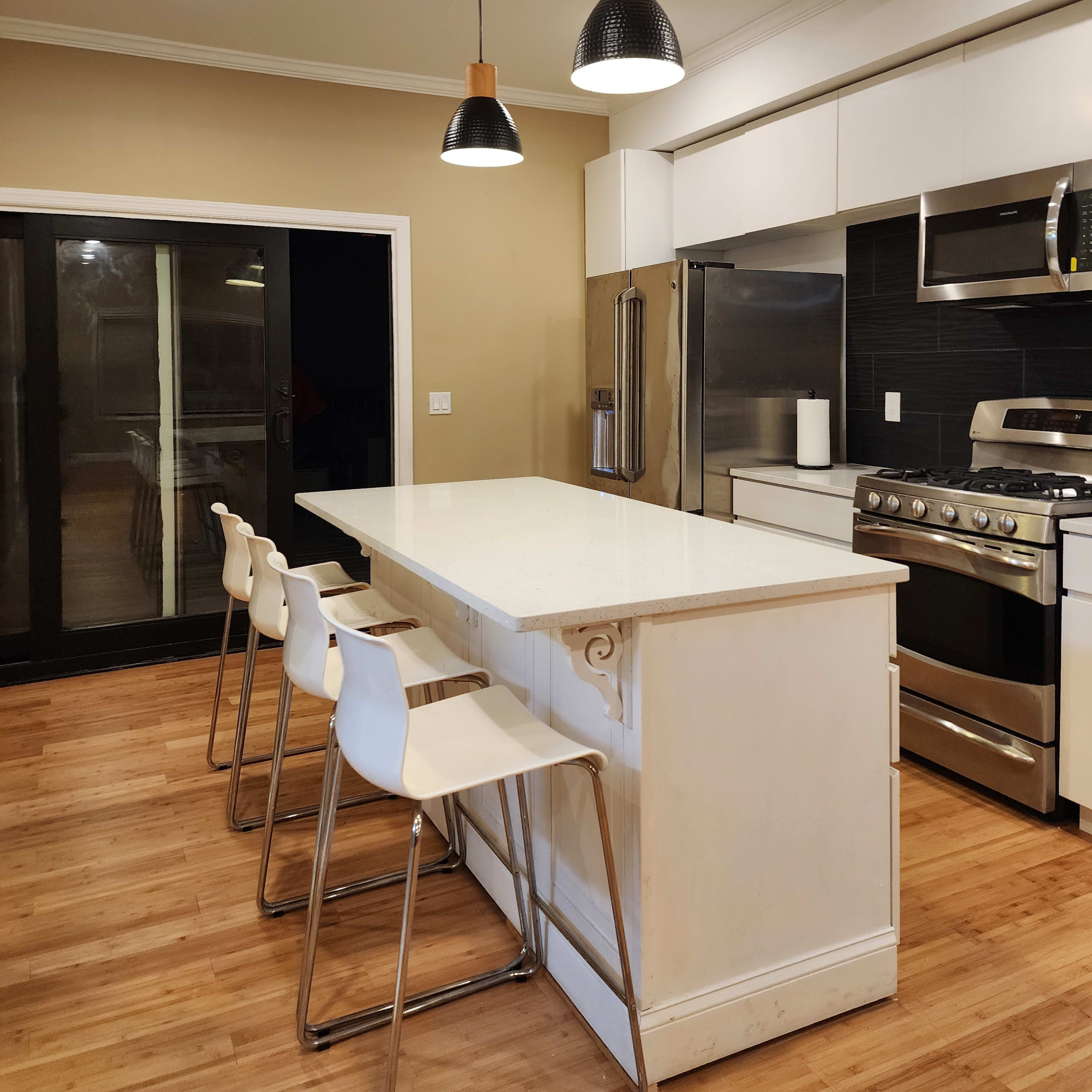 A modern kitchen features a large island with four white stools, stainless steel appliances, and three pendant lights overhead.