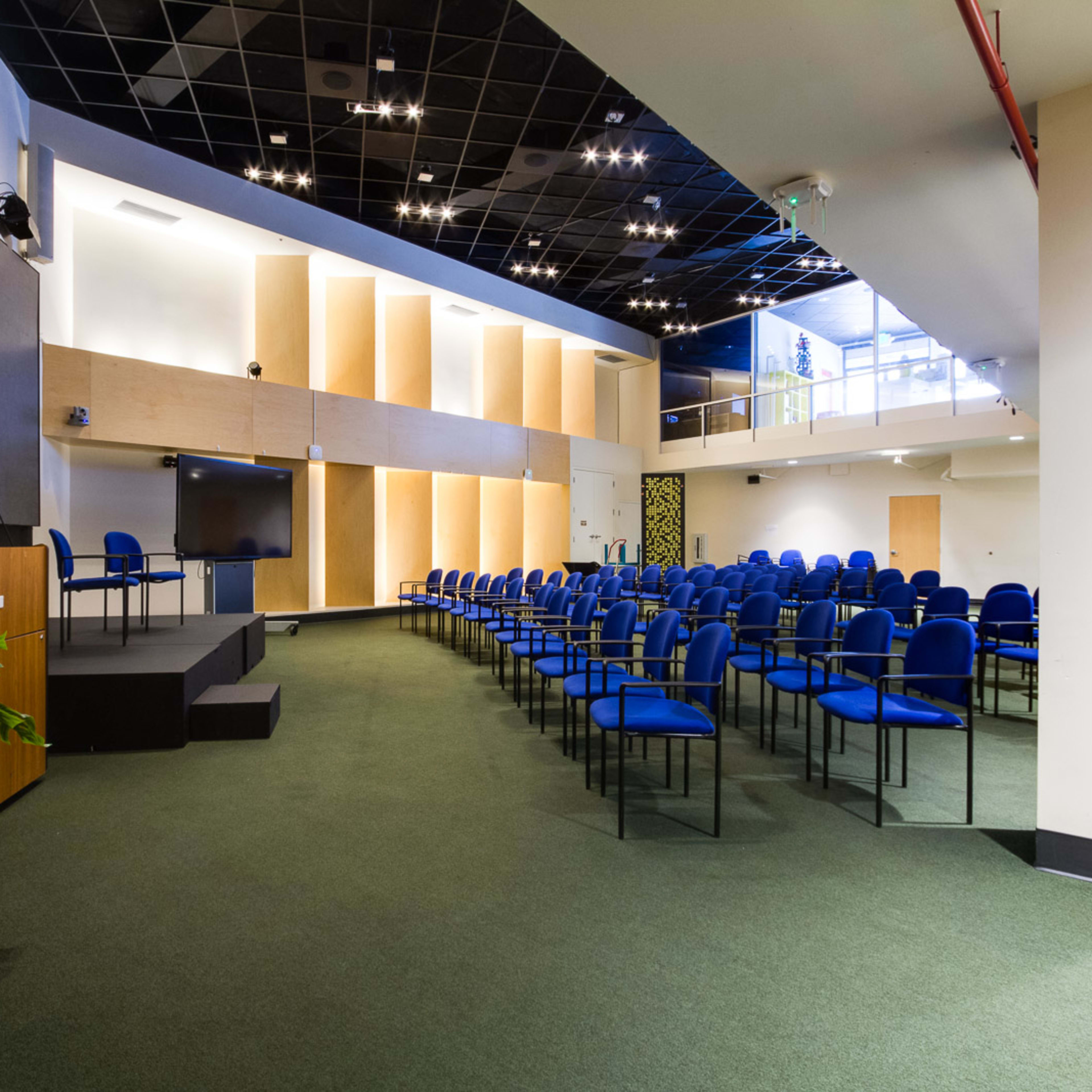 A spacious conference room with rows of blue chairs facing a podium, a screen, and a staircase leading to an upper level.