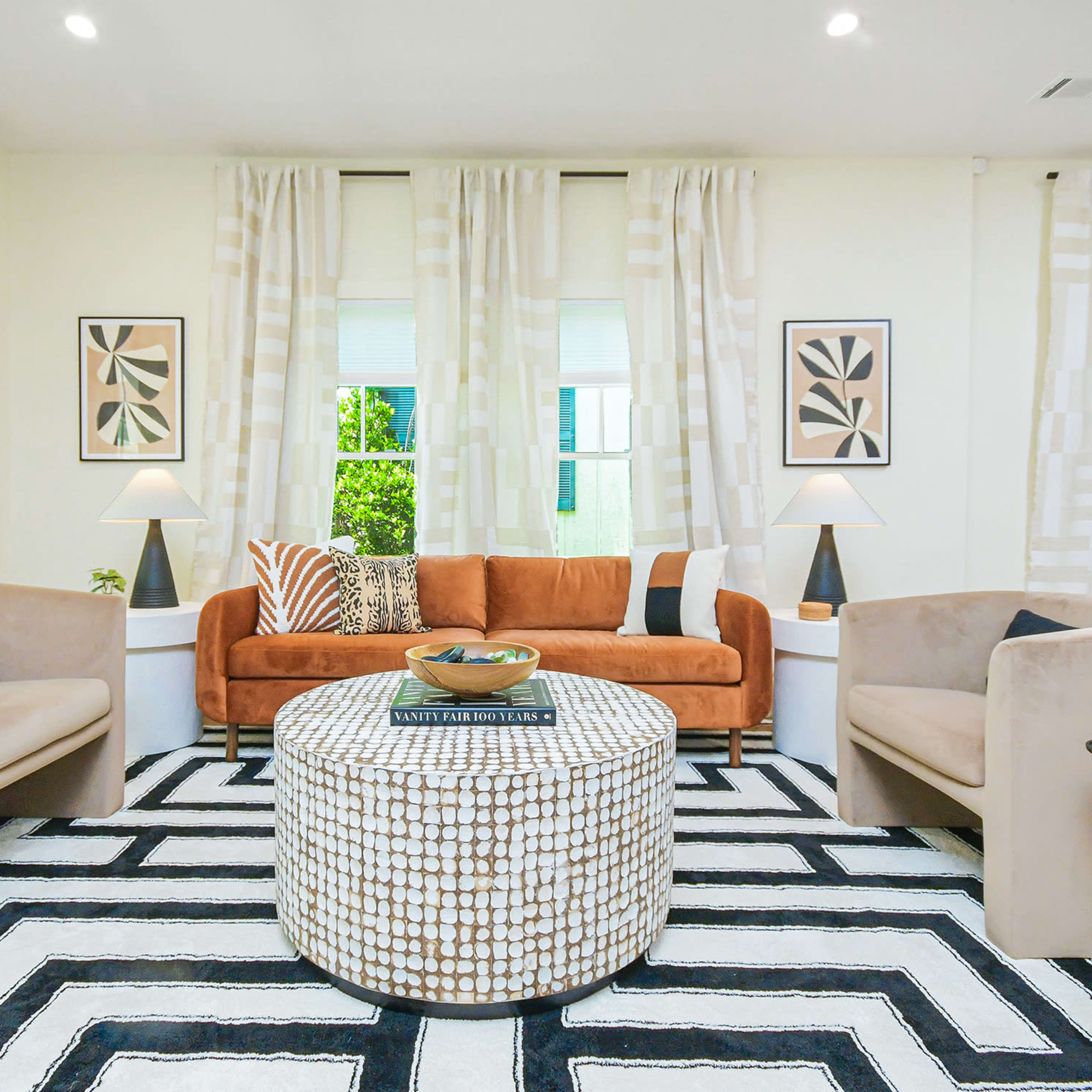 A well-furnished living room featuring a brown couch, two beige armchairs, a circular patterned coffee table, and large windows adorned with sheer curtains.