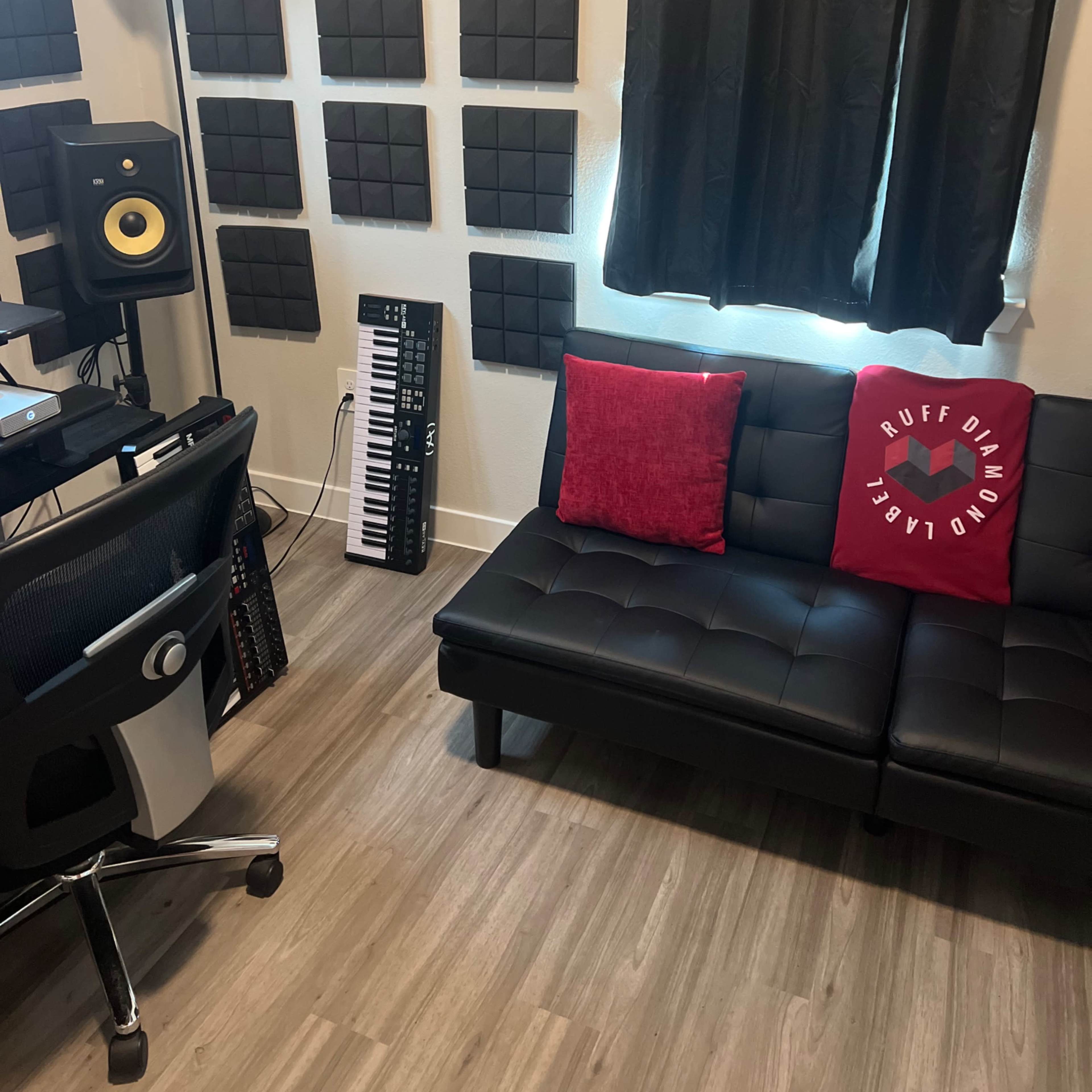 A music production studio featuring a black couch, a keyboard, acoustic panels on the walls, and a workstation with a lamp.
