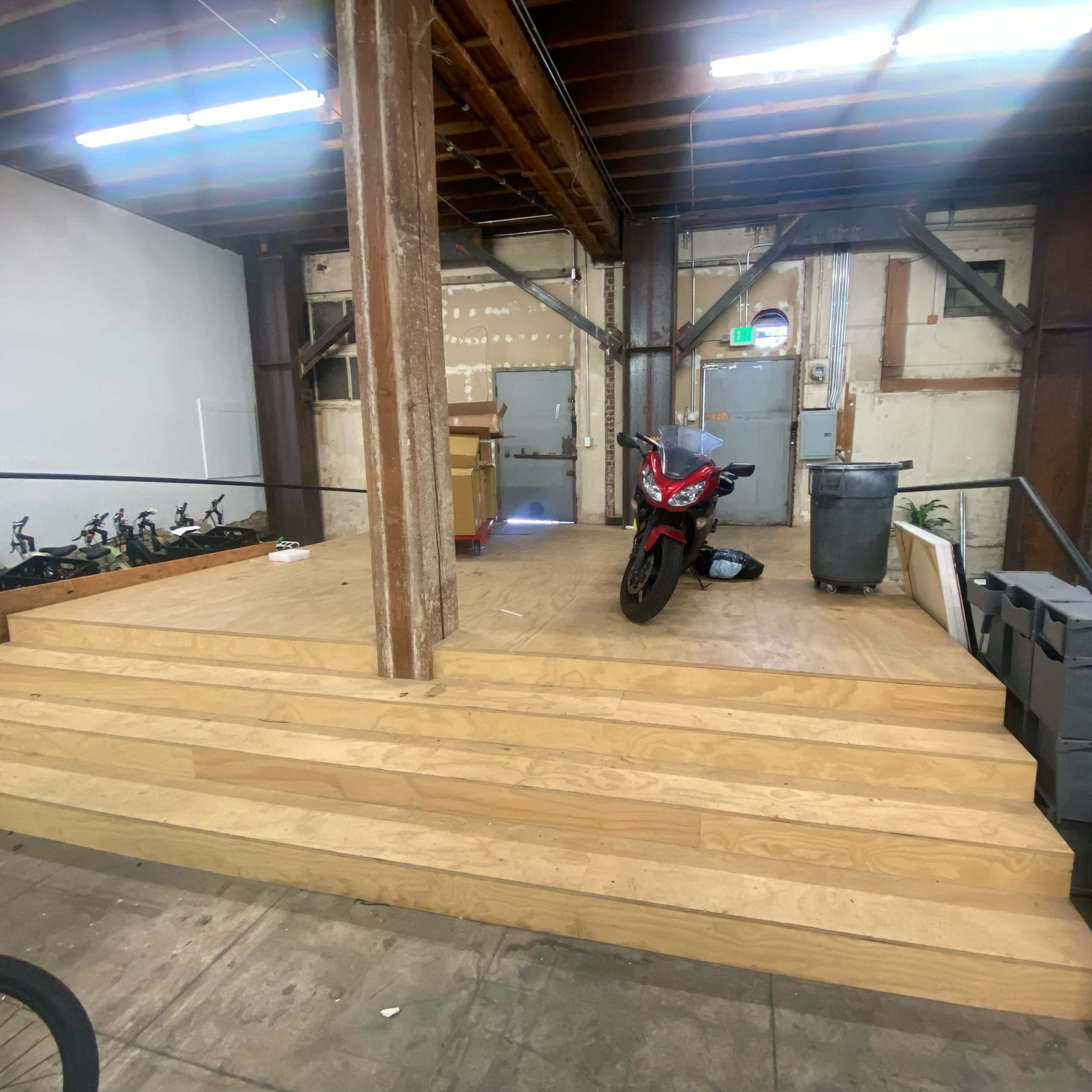 A wooden platform with steps, a motorcycle, and a trash can positioned against a wall in an industrial space.