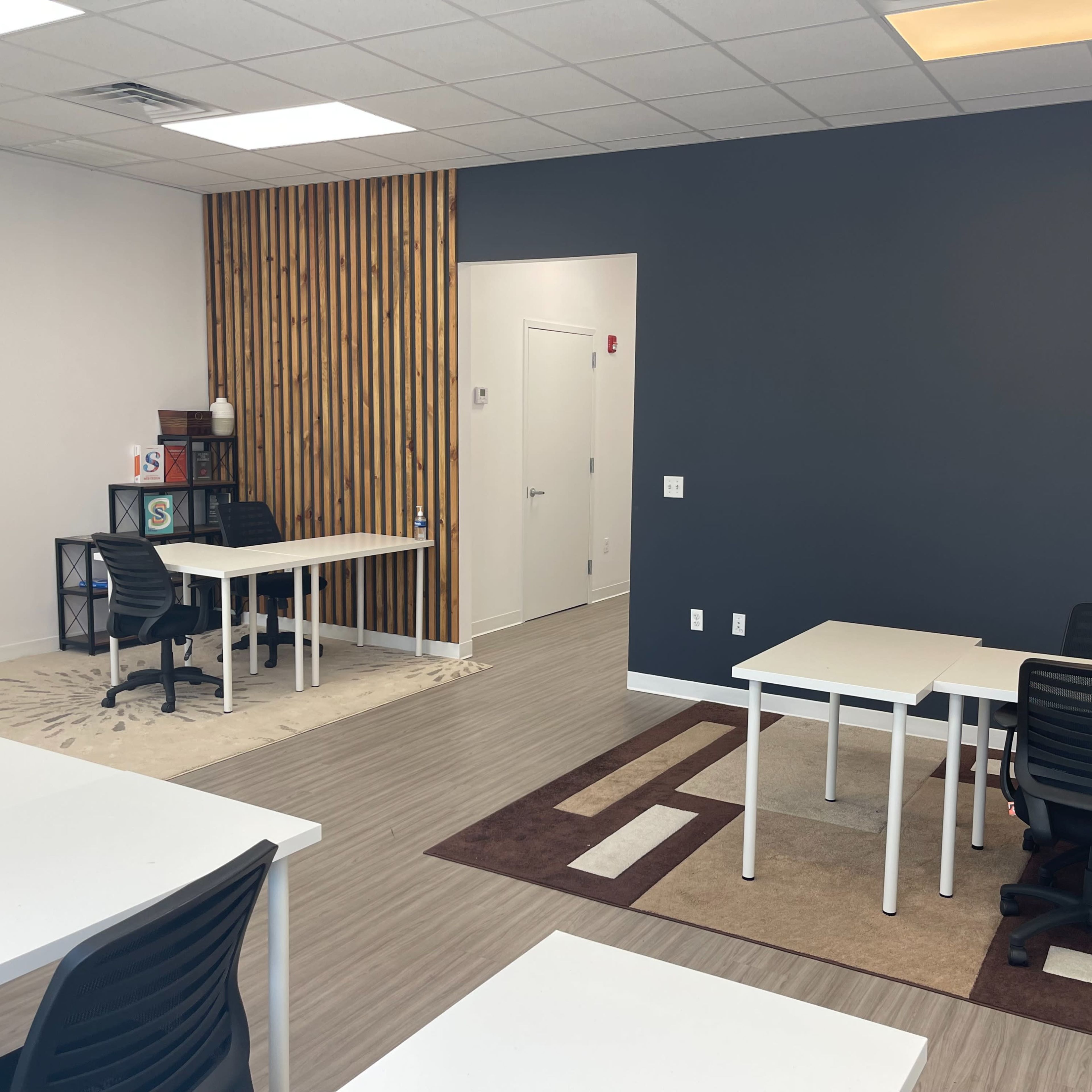 An office space with several white desks and black chairs, set against a backdrop of a dark gray wall and a wooden accent wall.