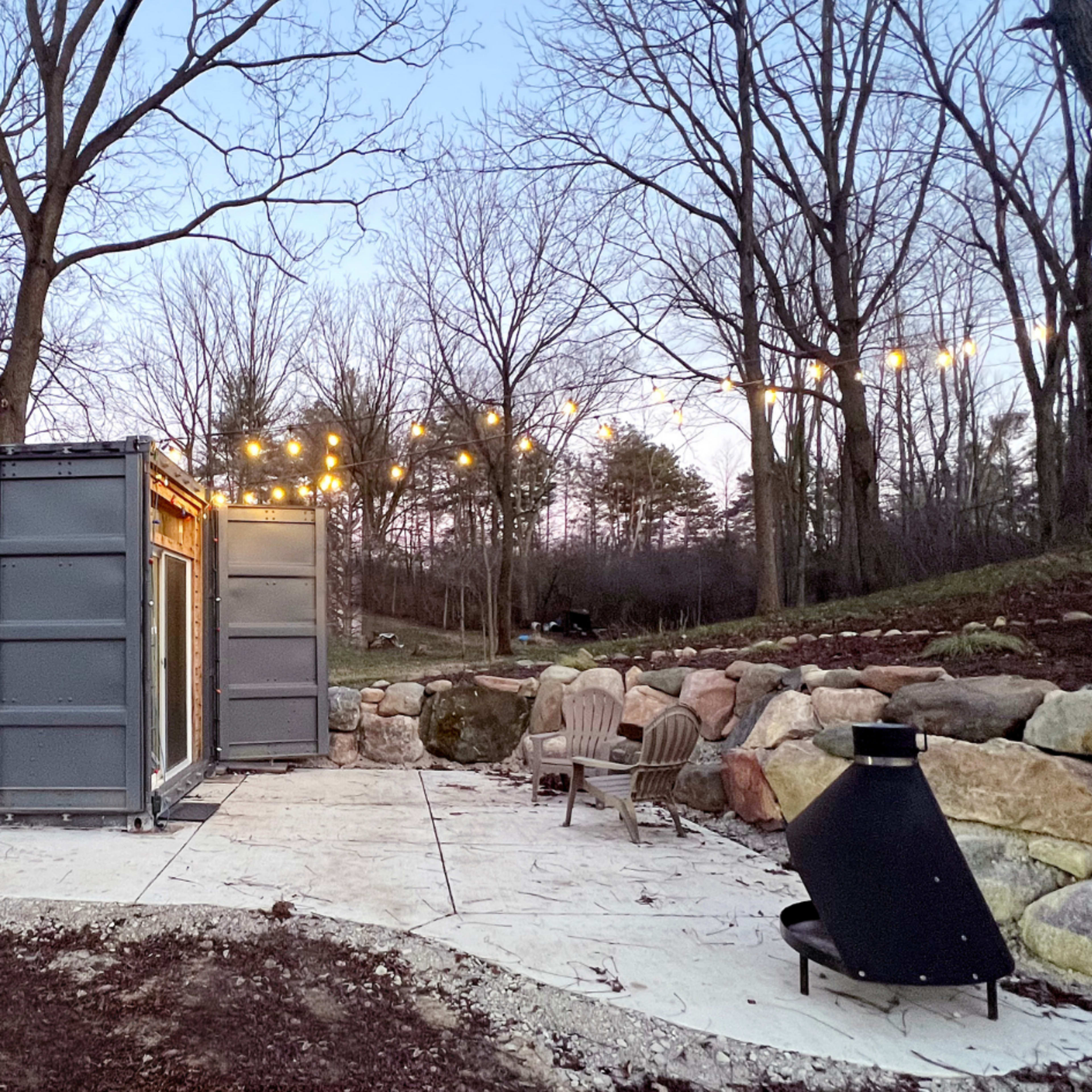A modern container home surrounded by trees, with patio lights strung overhead and a fire pit along a stone wall.