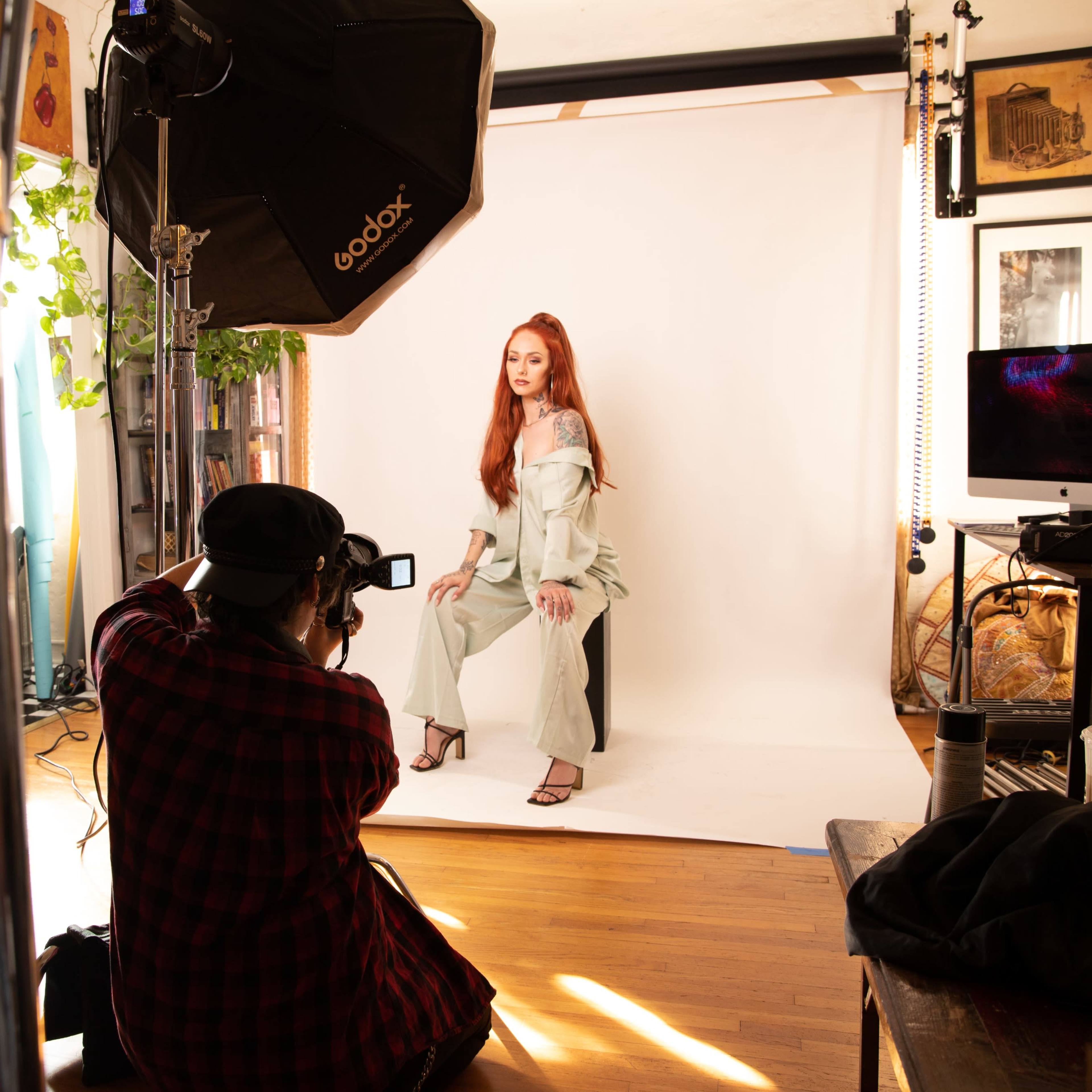 A photographer captures a model posing on a chair in a bright studio with a white backdrop and various equipment surrounding them.