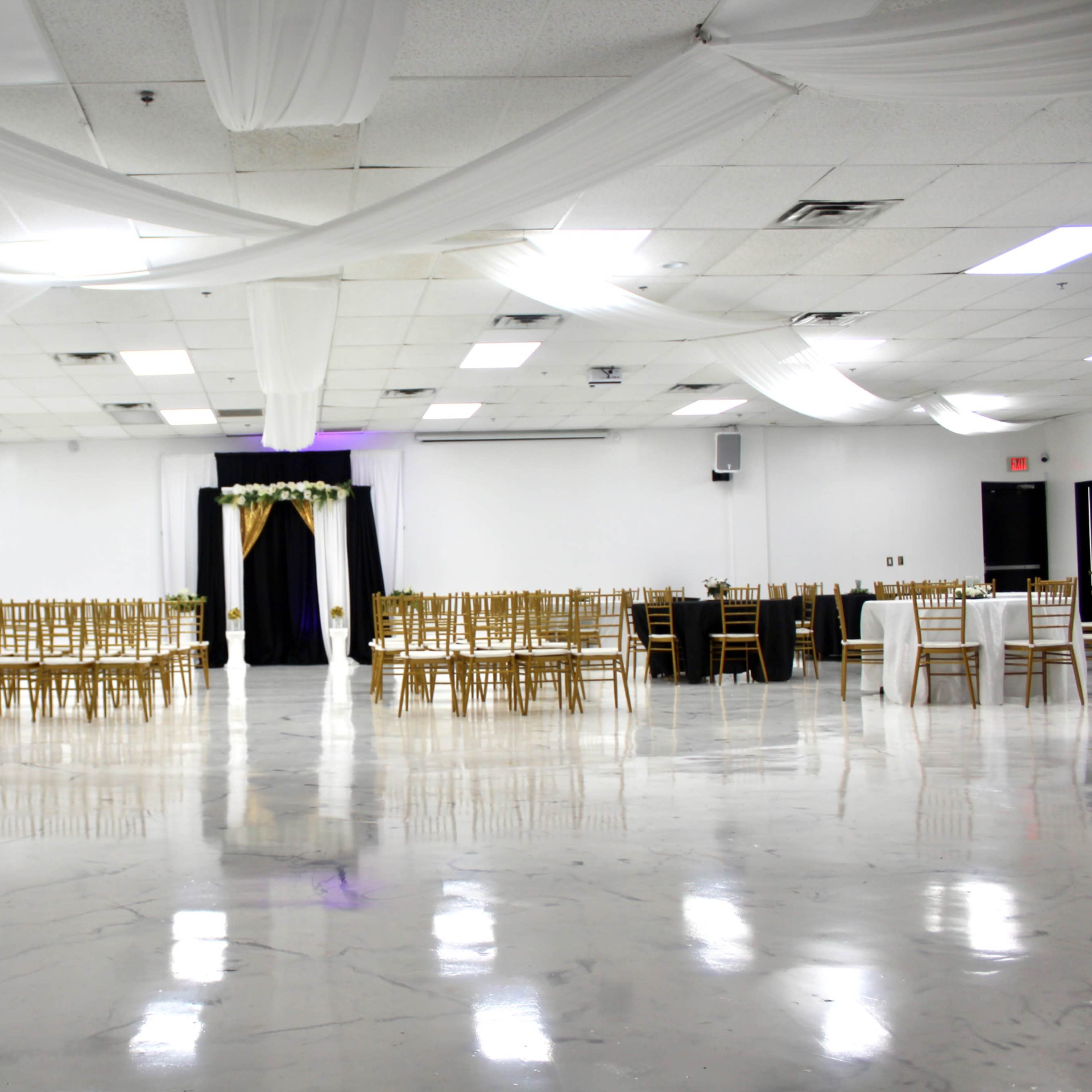 A spacious event hall features rows of gold chairs, tables set for dining, and decorative drapery above.