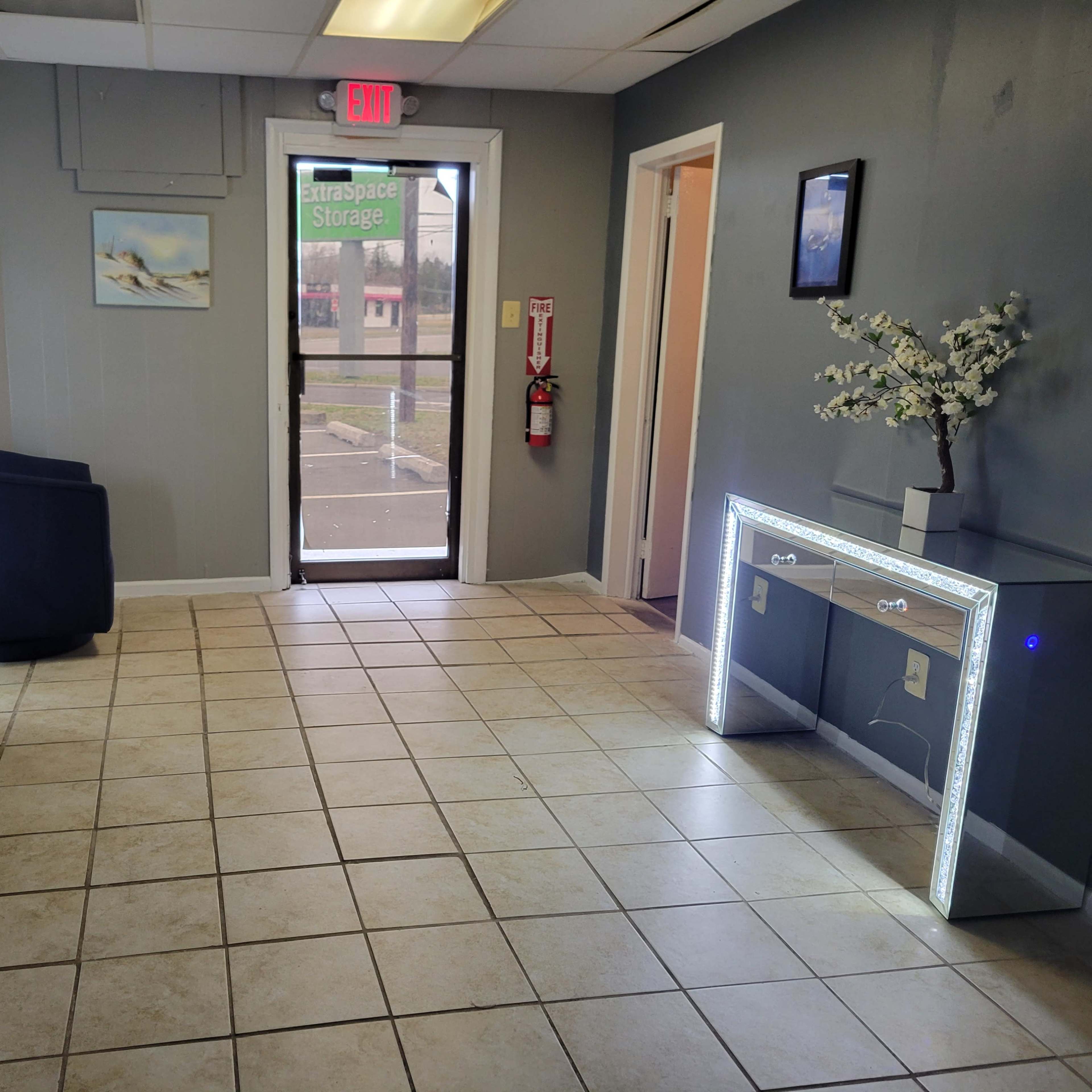 A small, well-lit reception area featuring a gray sofa, a blue chair, a glass console table with LED lights, and a door leading outside.