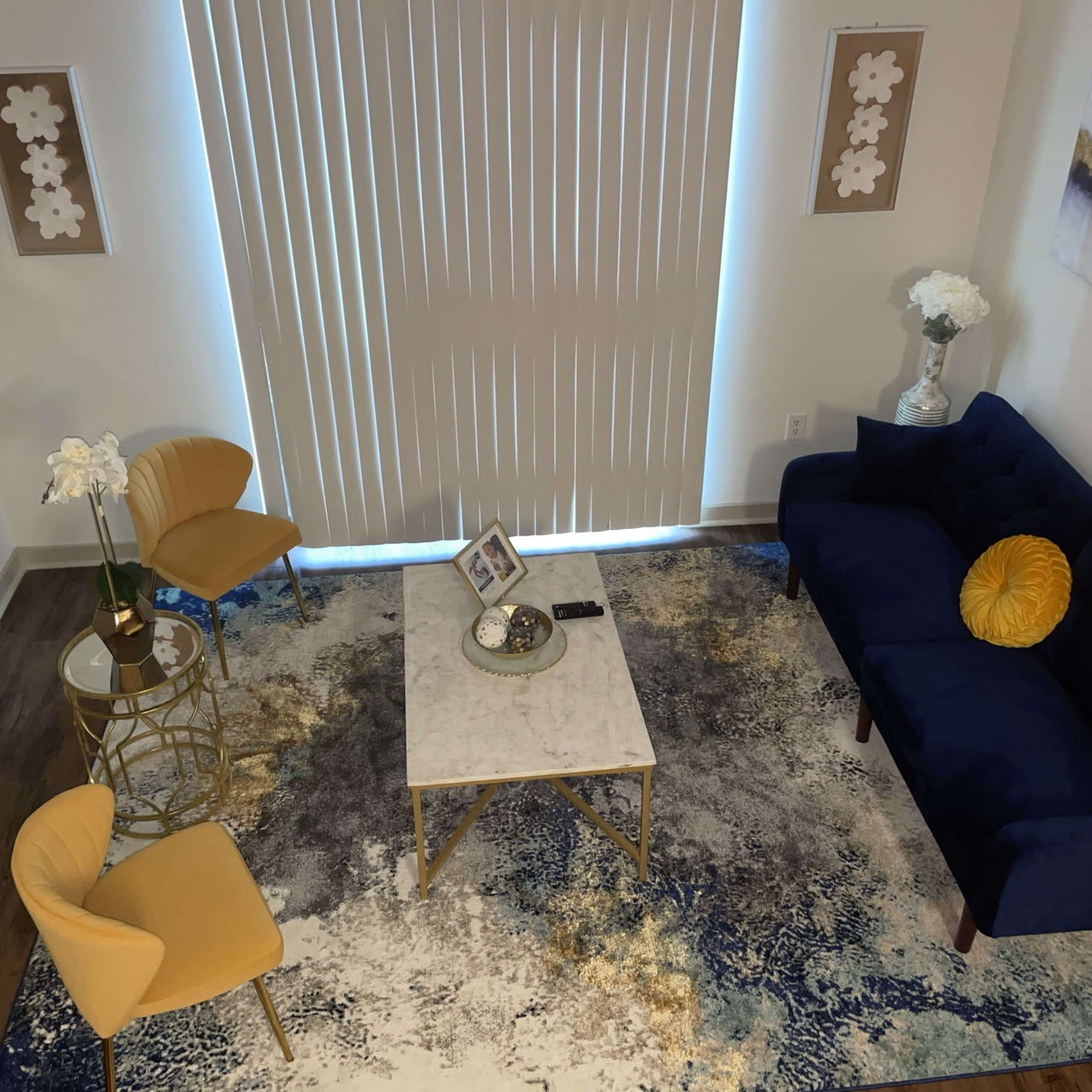 A cozy living room setup with a blue sofa, two yellow chairs, a marble coffee table, and decorative elements on a blue and white rug under vertical blinds.