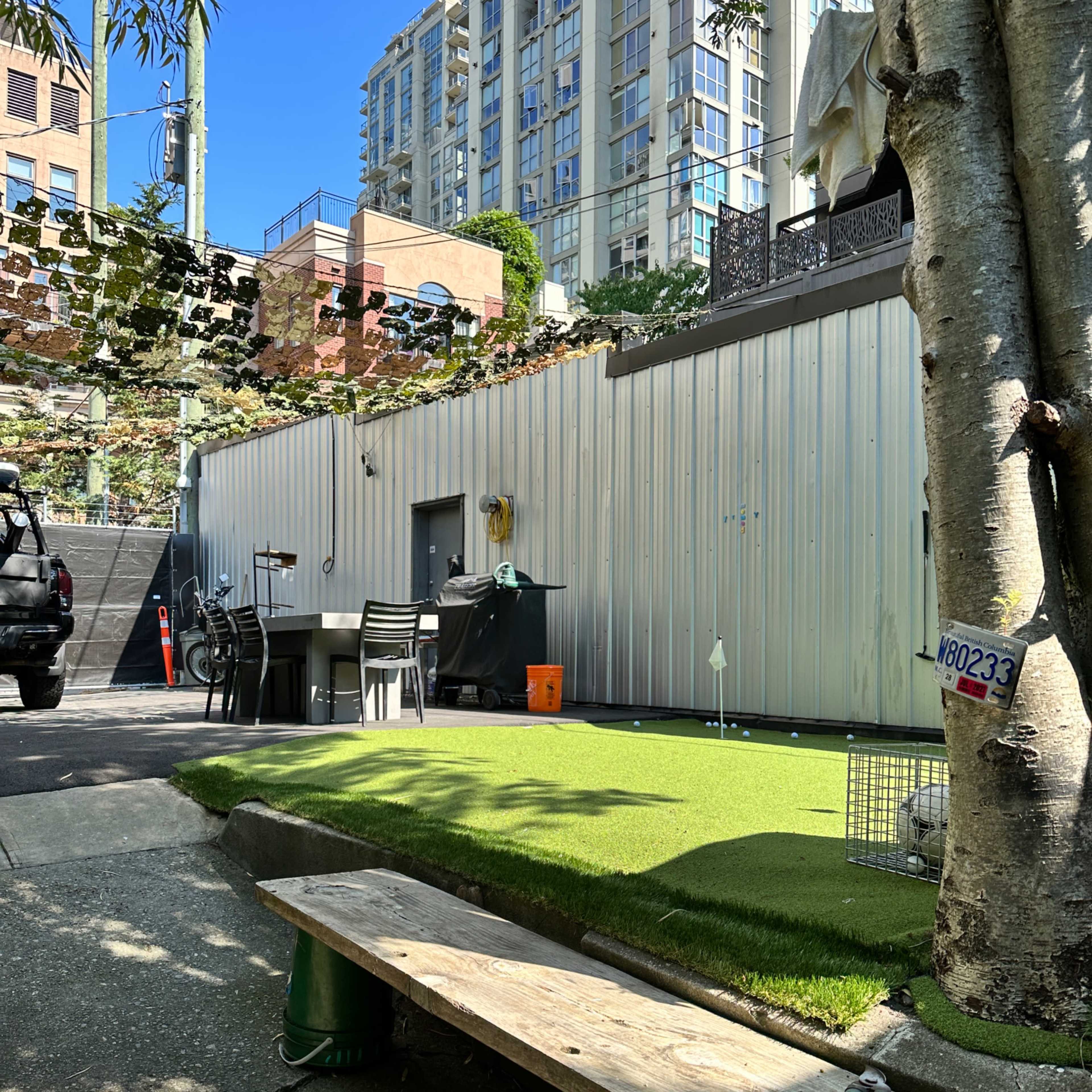 A small grassy area with a wooden bench and chairs in an urban setting, surrounded by buildings and a wall.