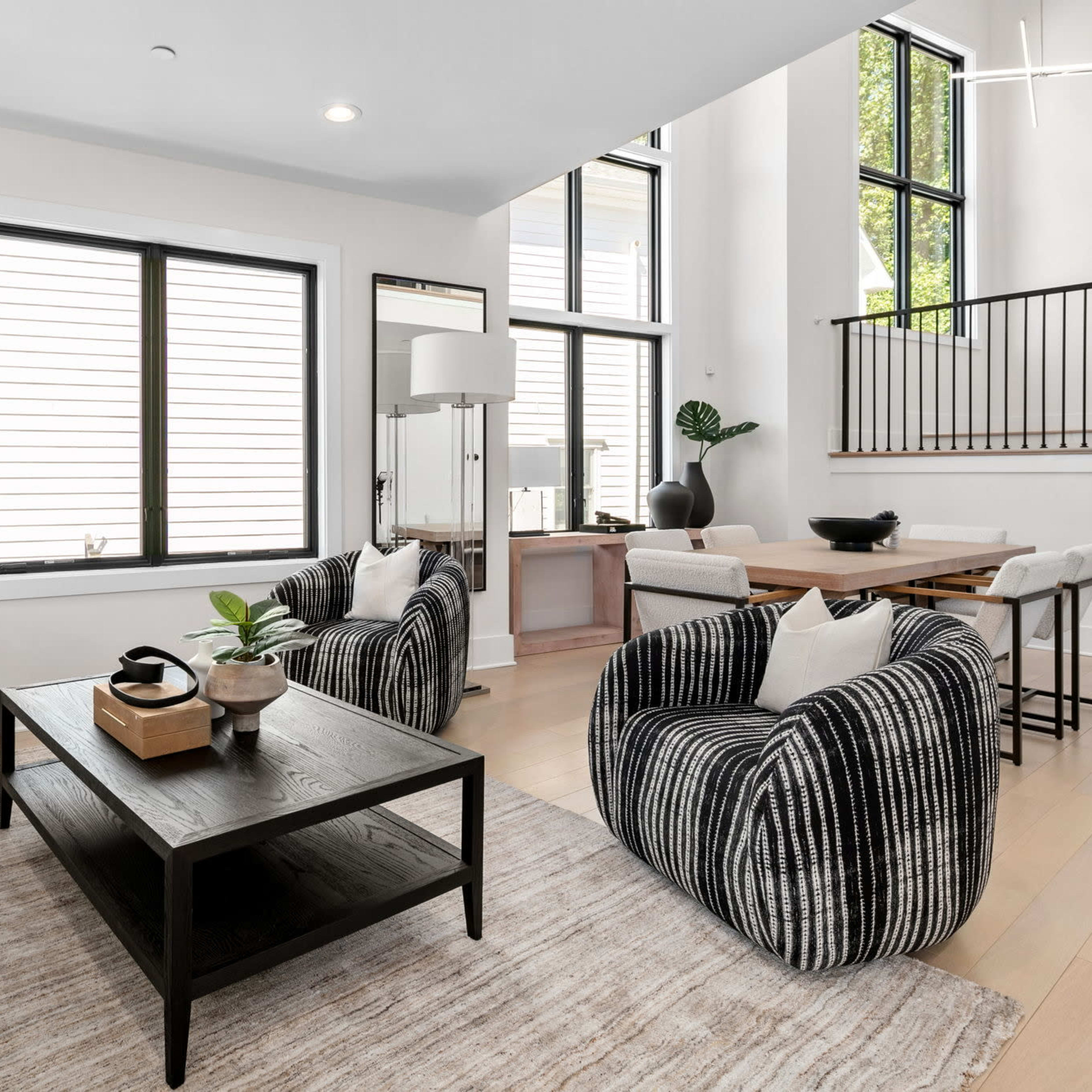 A modern living room features a beige sofa, striped armchairs, a dark coffee table, and large windows allowing natural light to enter.
