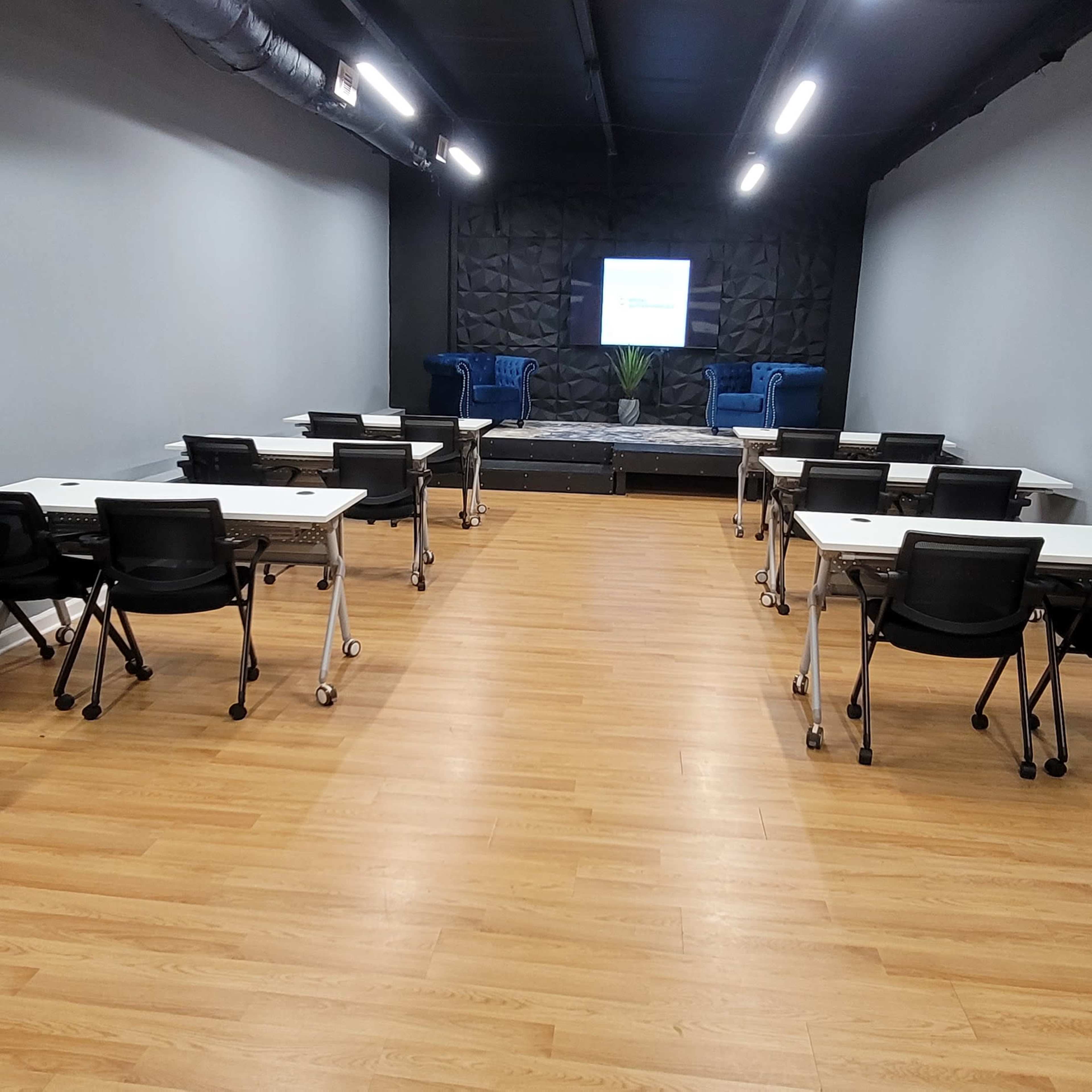The room features several tables and chairs arranged in rows facing a screen at the front, with a seating area in the background.