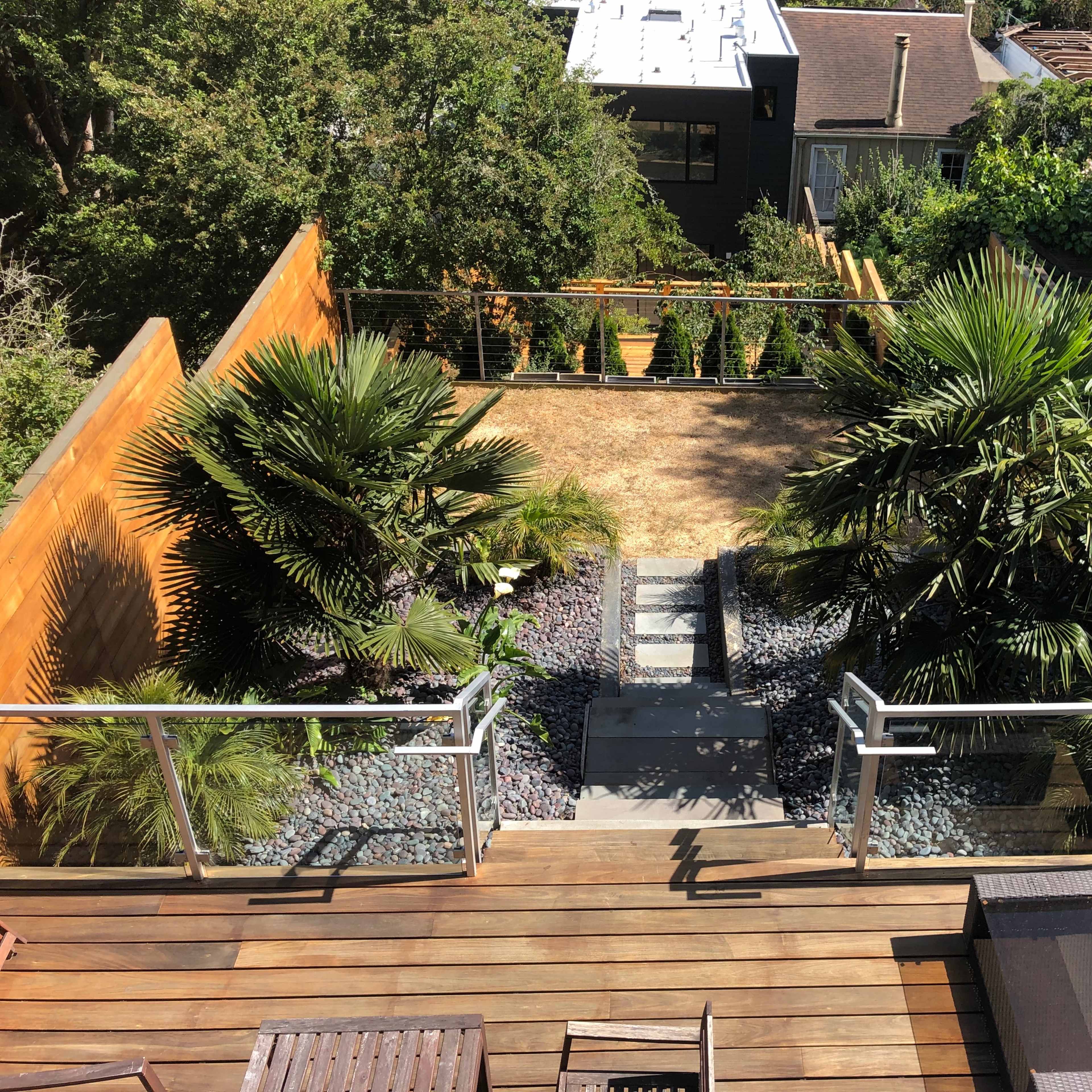 A backyard with a stone pathway leading down to an area featuring two palm trees and gravel, enclosed by wooden fencing.