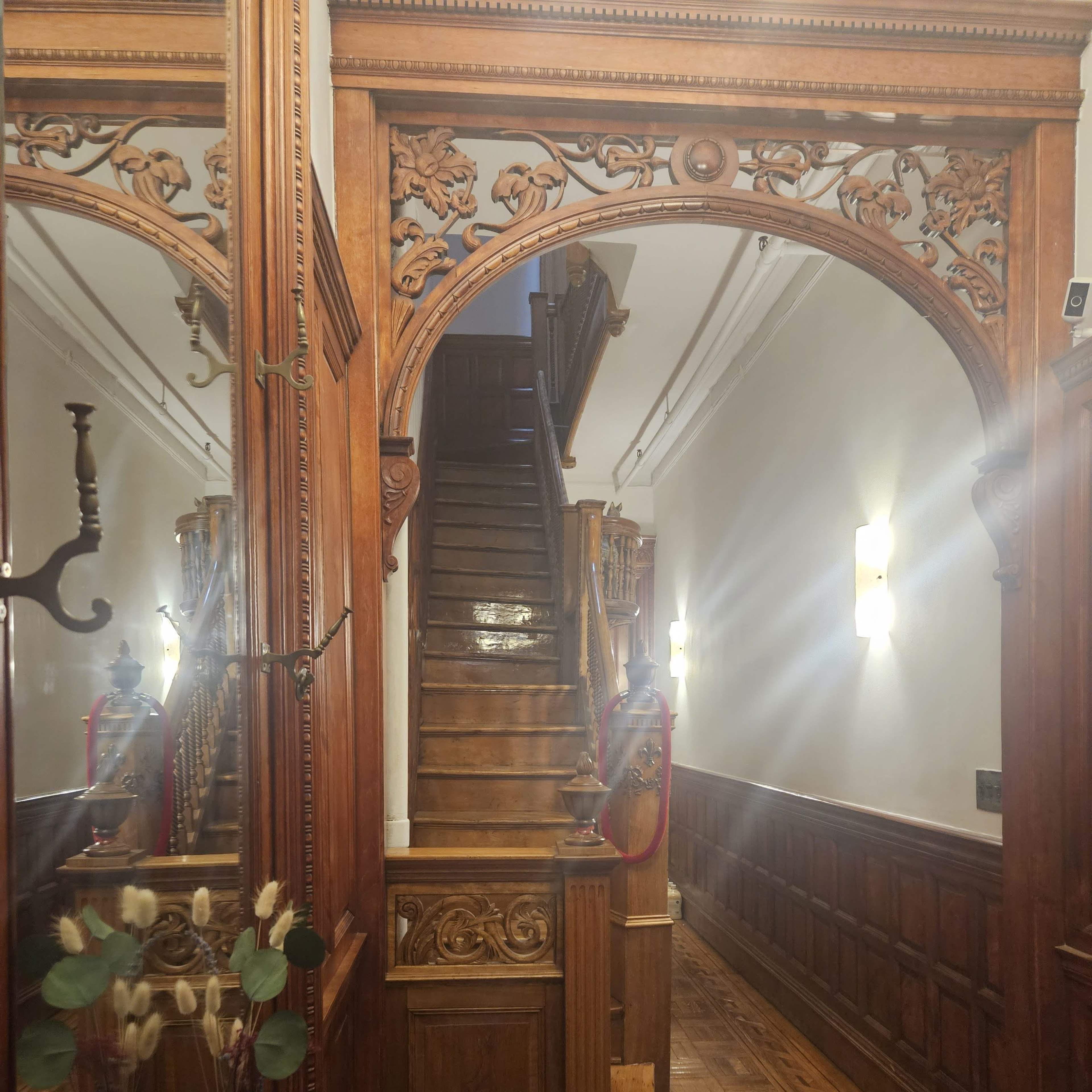 A wooden staircase leading to an upper level, framed by intricately carved wooden archways in a hallway with polished wooden flooring and wall-mounted lighting.