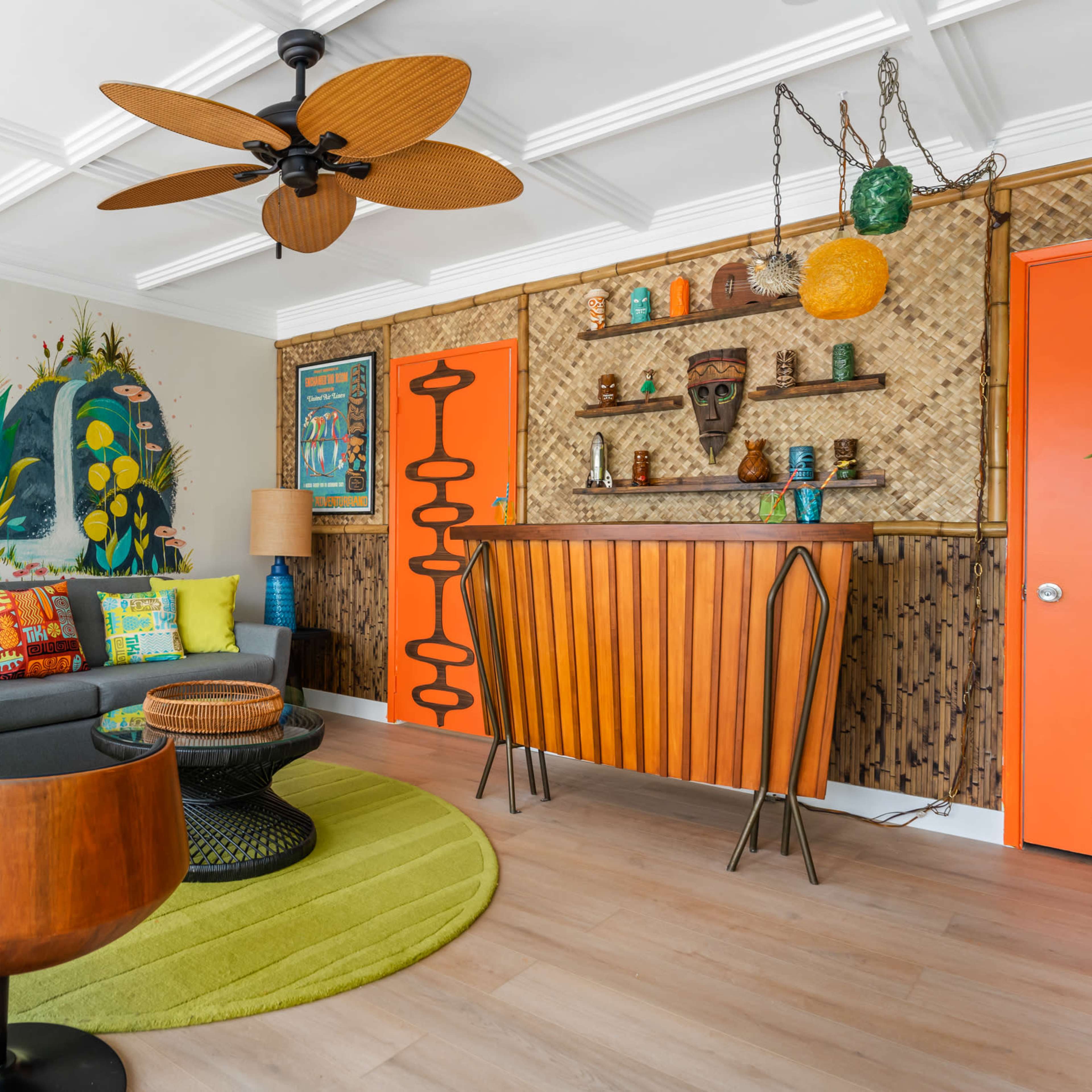 The living room features a colorful mural, a modern bar area with vibrant decorations, a round green rug, and wooden accent furniture.