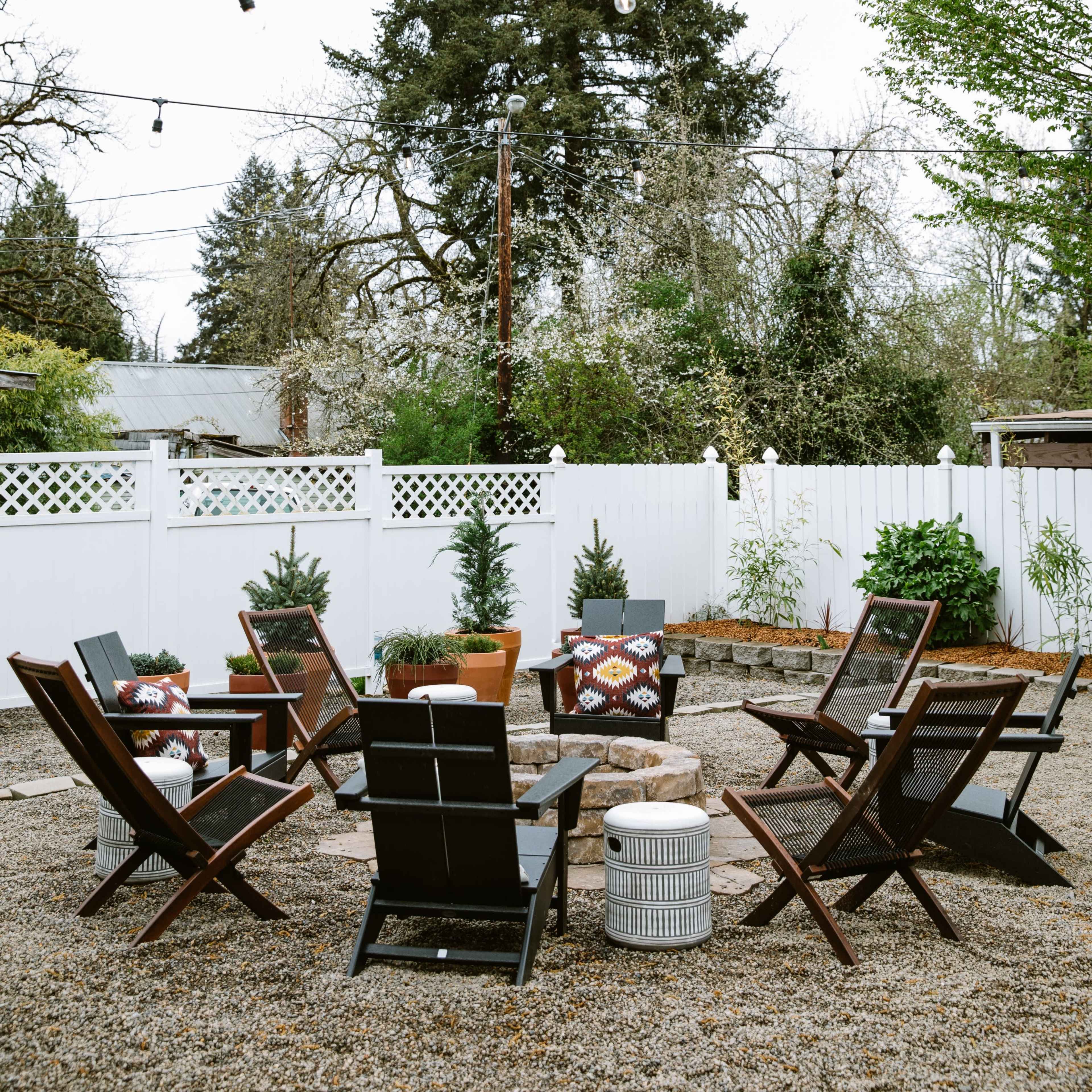 A gravel patio features several black chairs arranged around a central fire pit, surrounded by potted plants and a white picket fence.