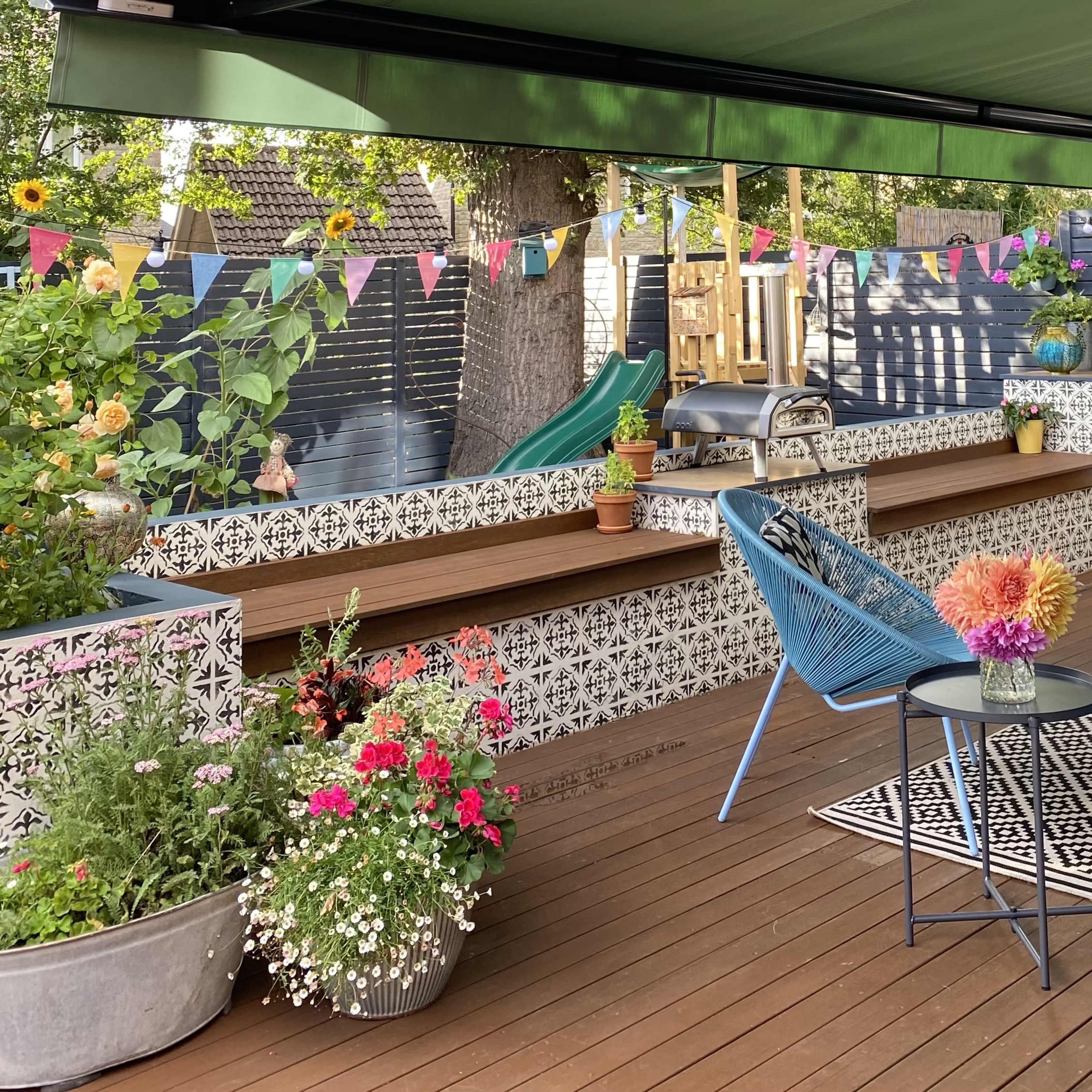 A spacious outdoor patio features blue seating, colorful flower arrangements, and a decorative banner, set among wooden decking and patterned tile.