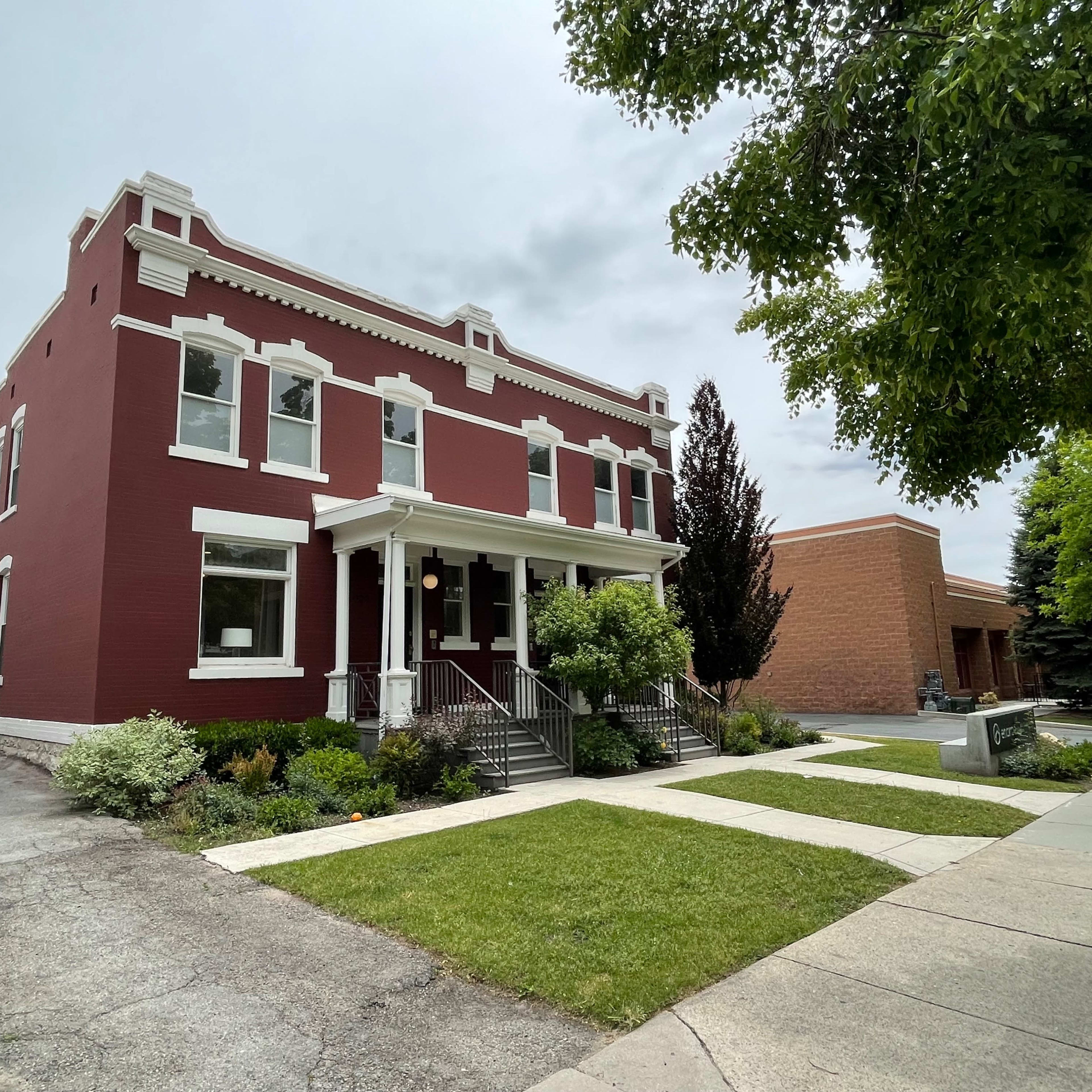 A red-brick building with white trim, featuring a front porch and landscaped greenery, situated on a sidewalk beside a paved road.