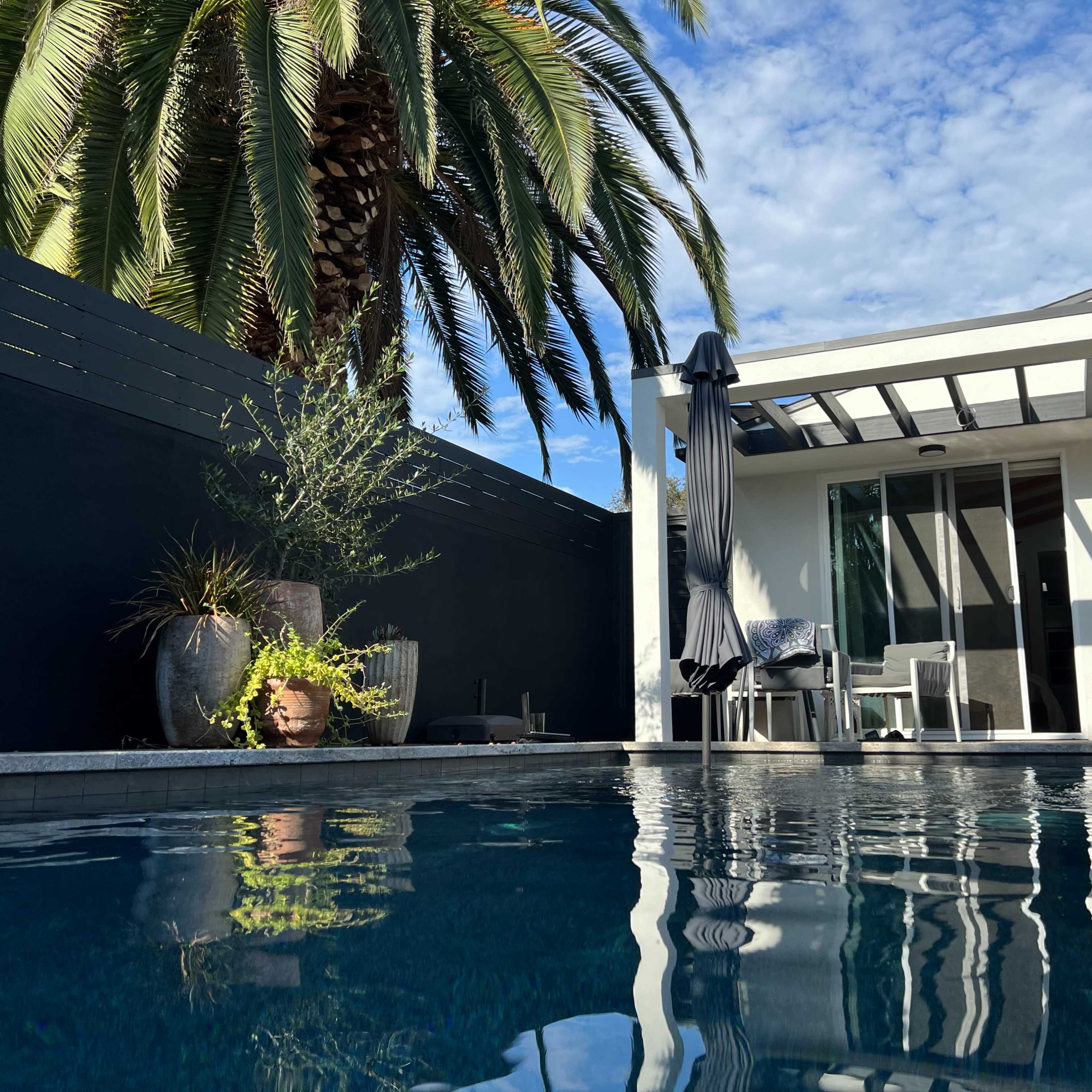 A modern poolside area featuring a minimalist structure, palm trees, and patio furniture.