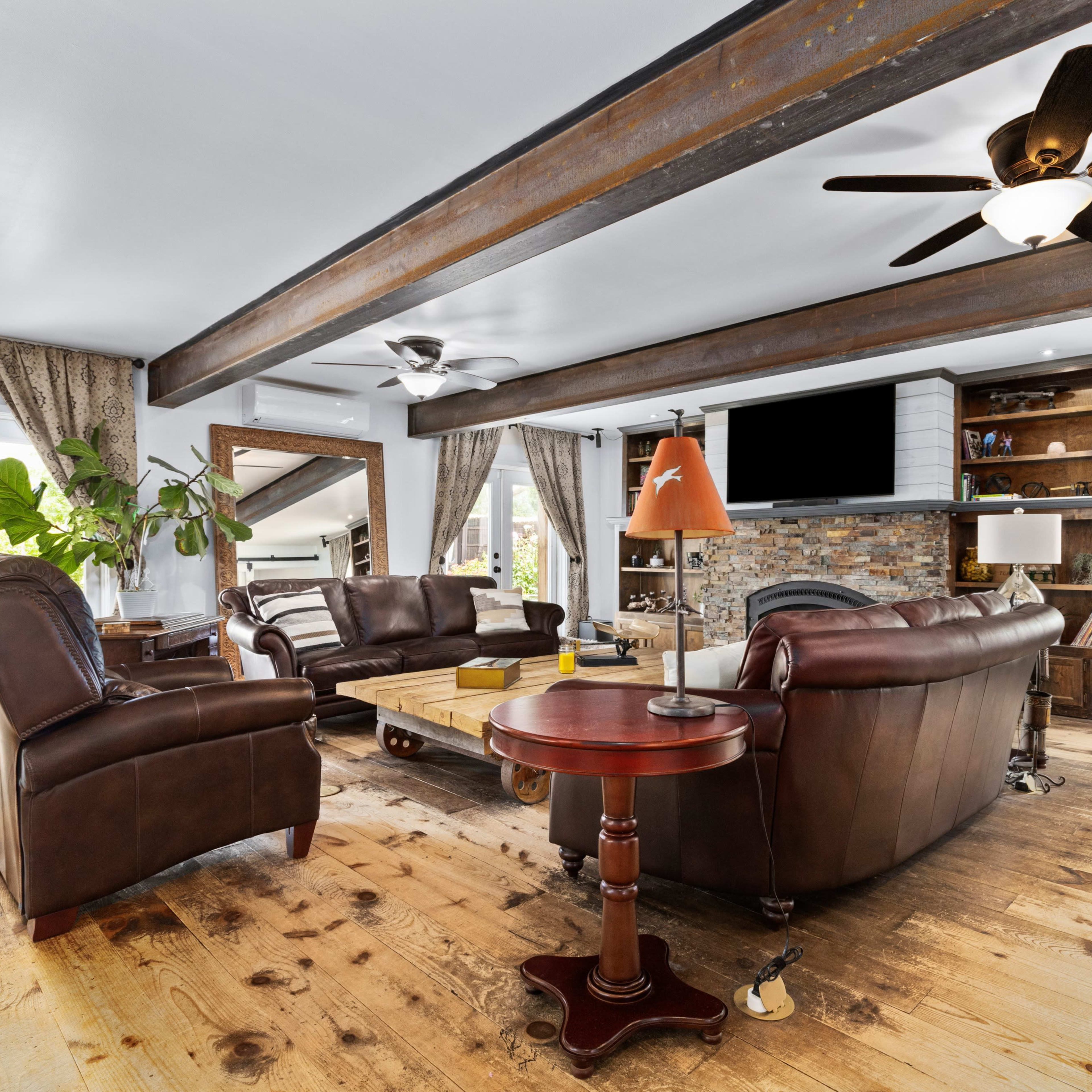 The living room features two leather sofas, a coffee table, a side table, and a stone fireplace, with wood beams overhead and large windows letting in natural light.