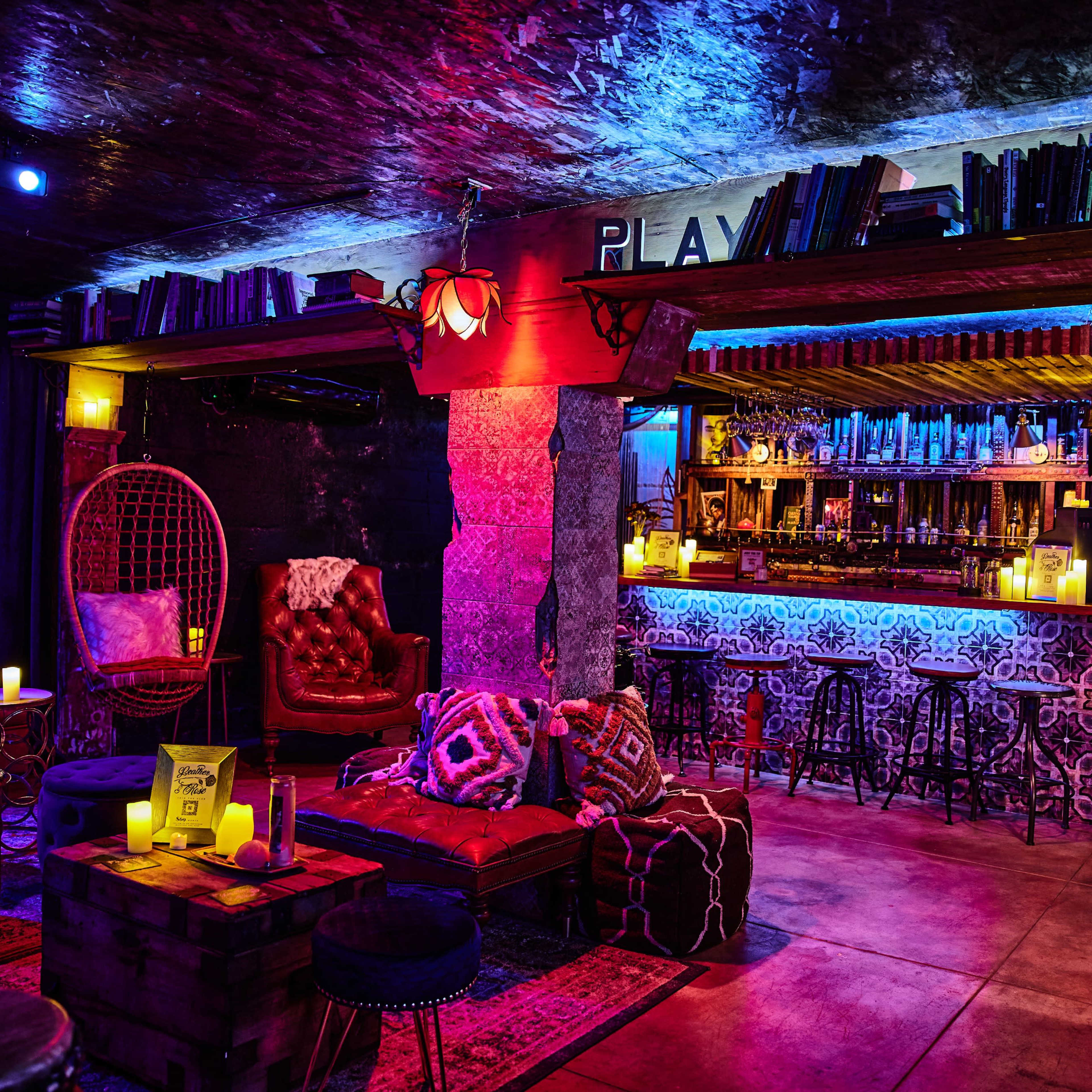The scene features a vibrant lounge area with eclectic furniture, a stocked bar, and decorative shelves filled with books, illuminated by colorful lights.