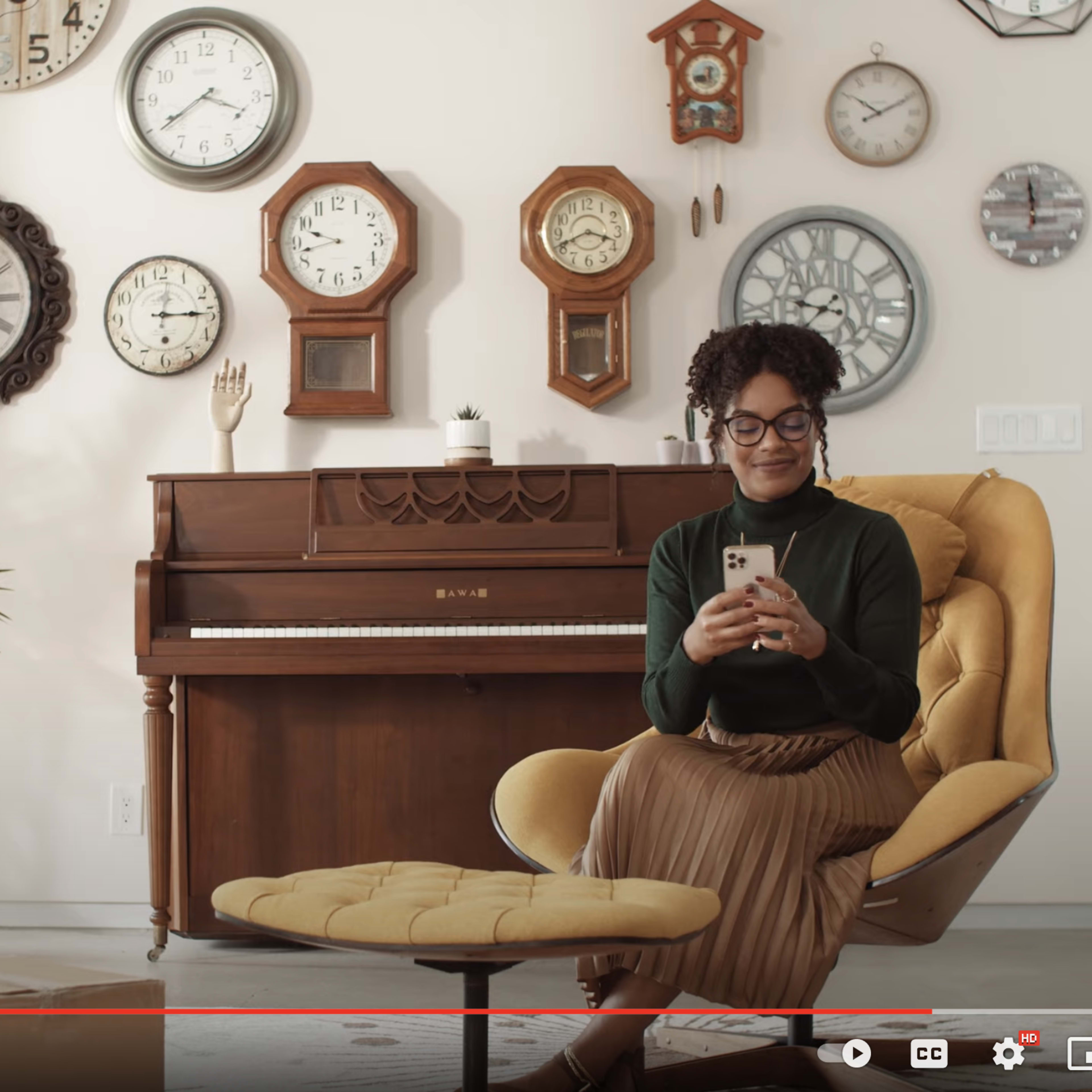 A woman sits on a yellow chair, using her phone, in a room adorned with various wall clocks and a wooden piano.