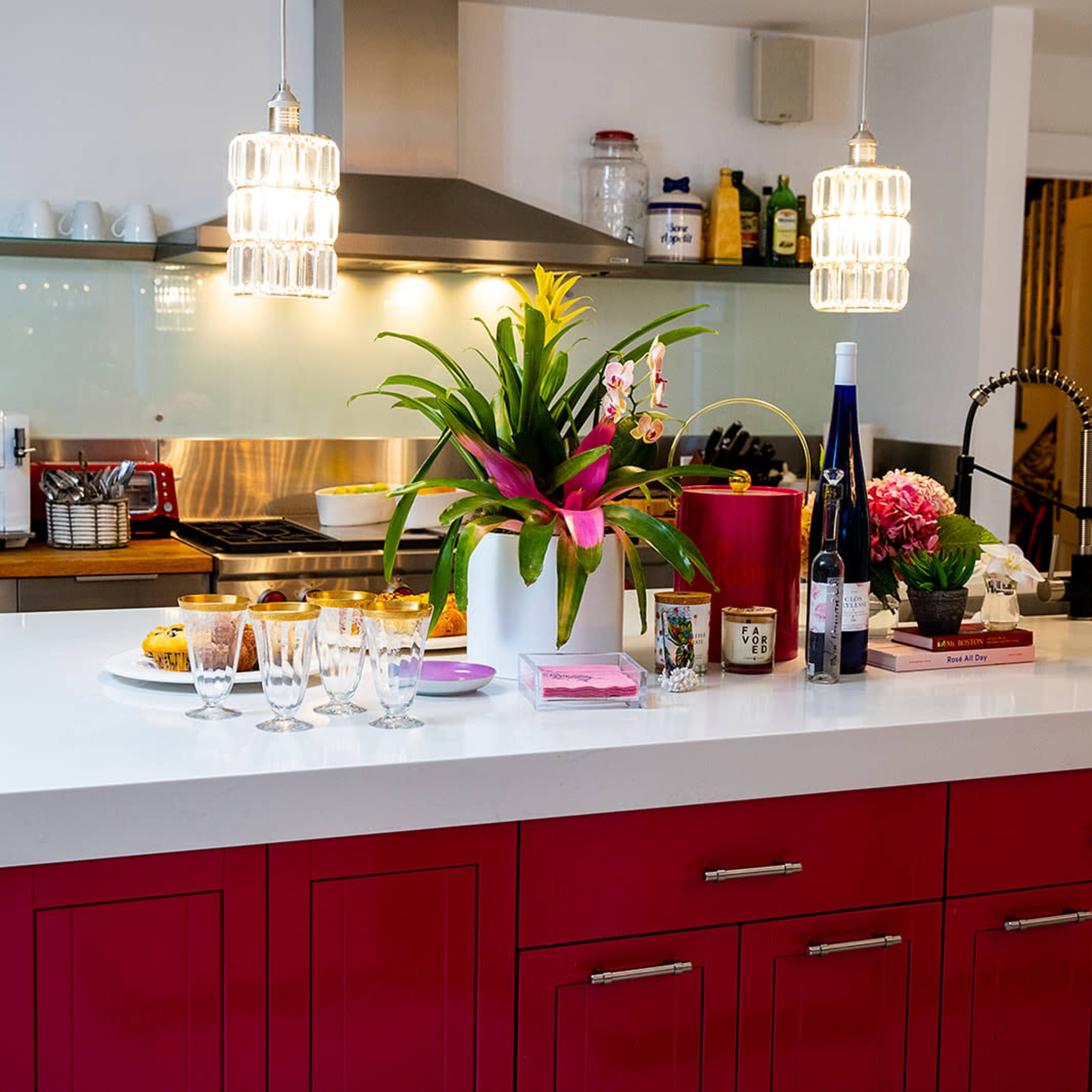 A modern kitchen features a bright red island with a countertop adorned with snacks, drinks, and a decorative plant under pendant lighting.