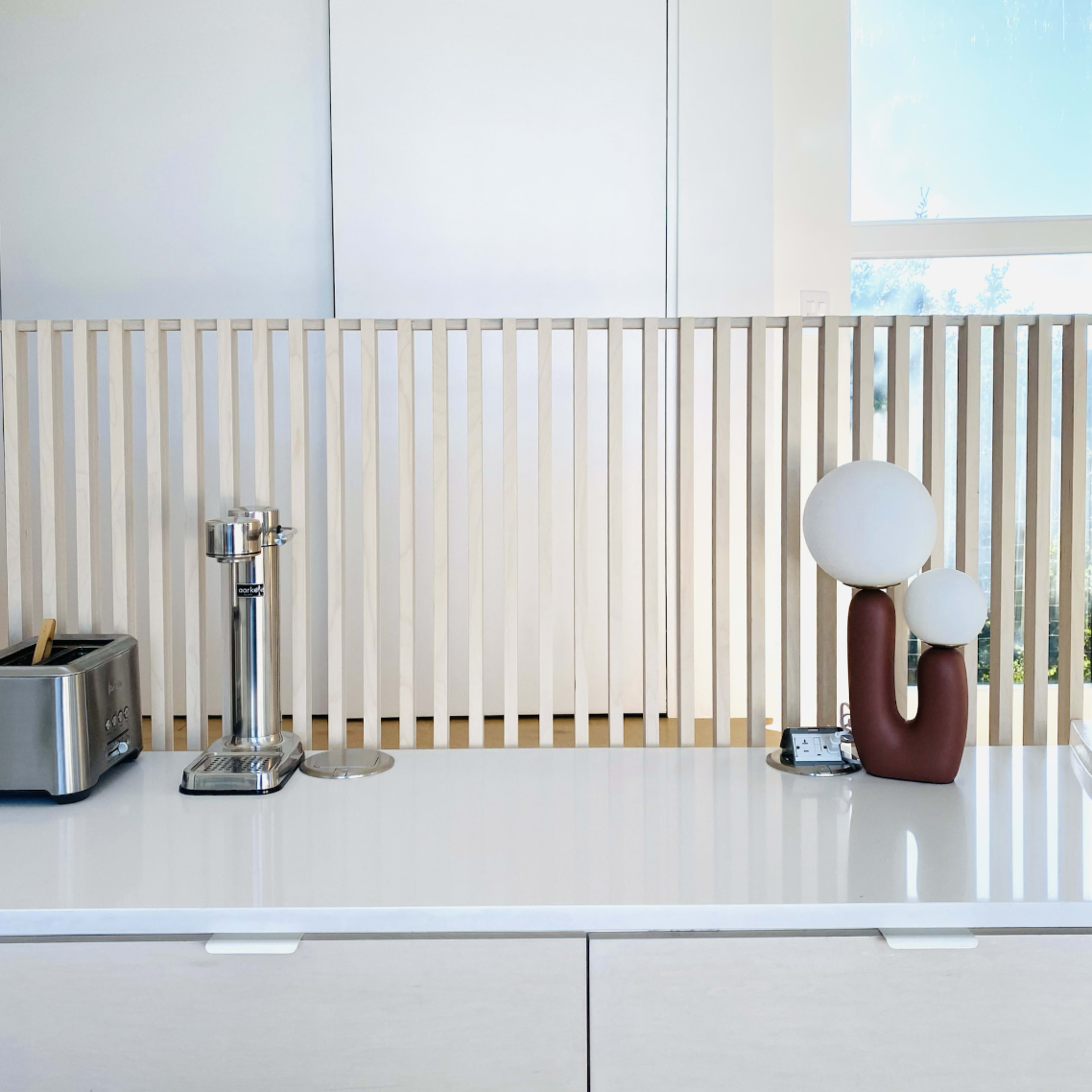 A sleek kitchen countertop features a toaster, a beverage tap, and a decorative lamp behind a wooden slatted partition.