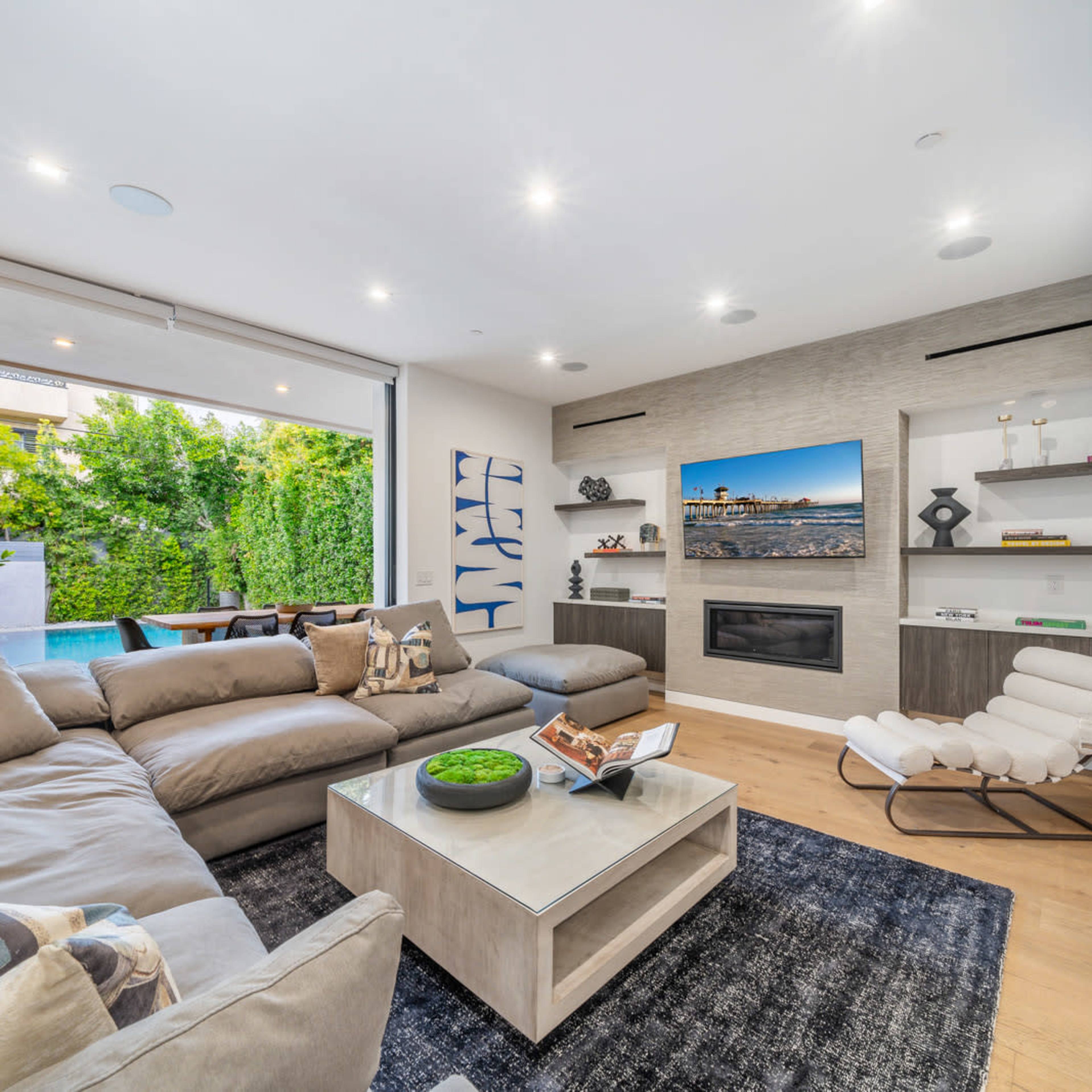 A modern living room features a large sectional sofa, a coffee table, and a wall-mounted television, with sliding glass doors leading to an outdoor patio.