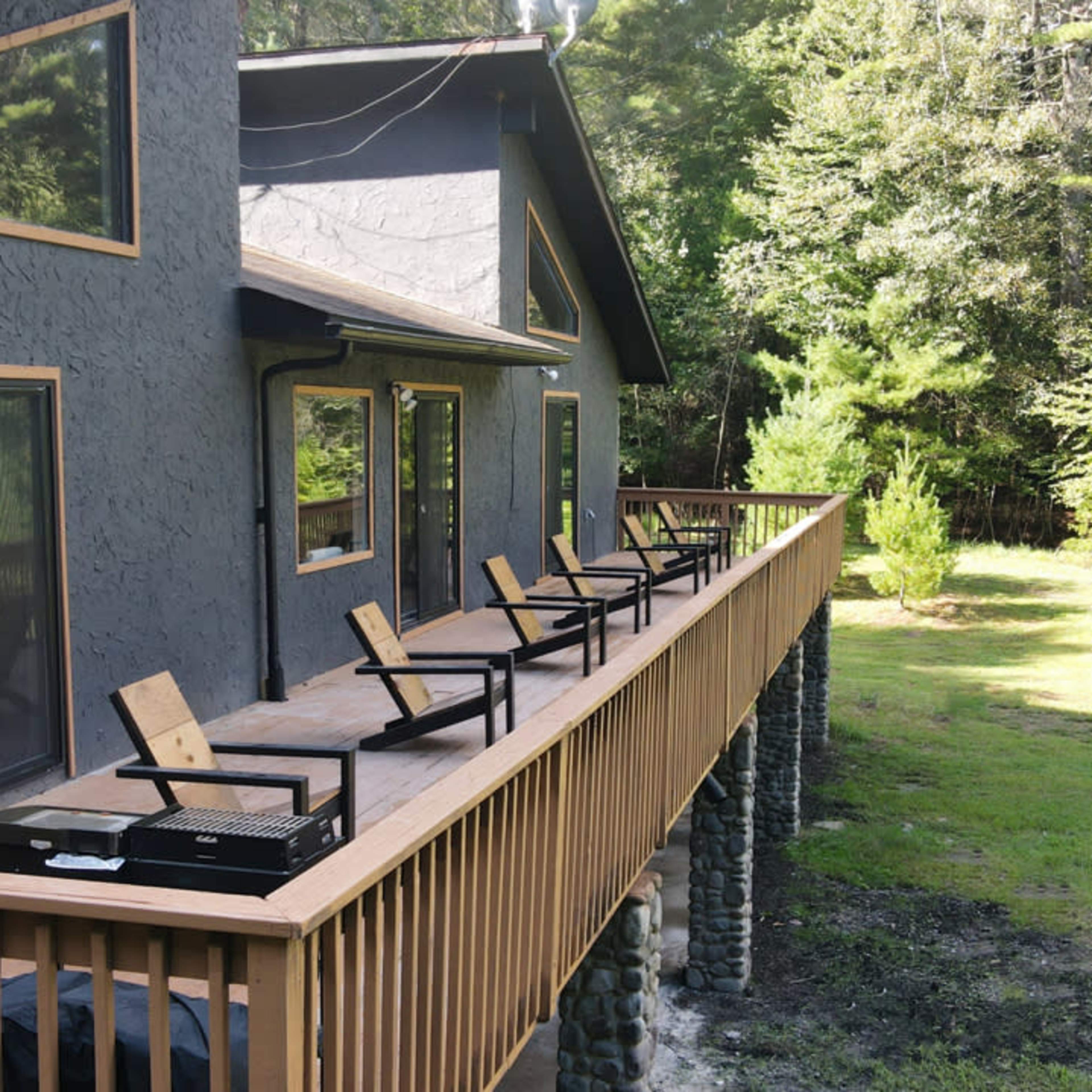 A wooden deck with several lounge chairs extends along the side of a house surrounded by tall trees.